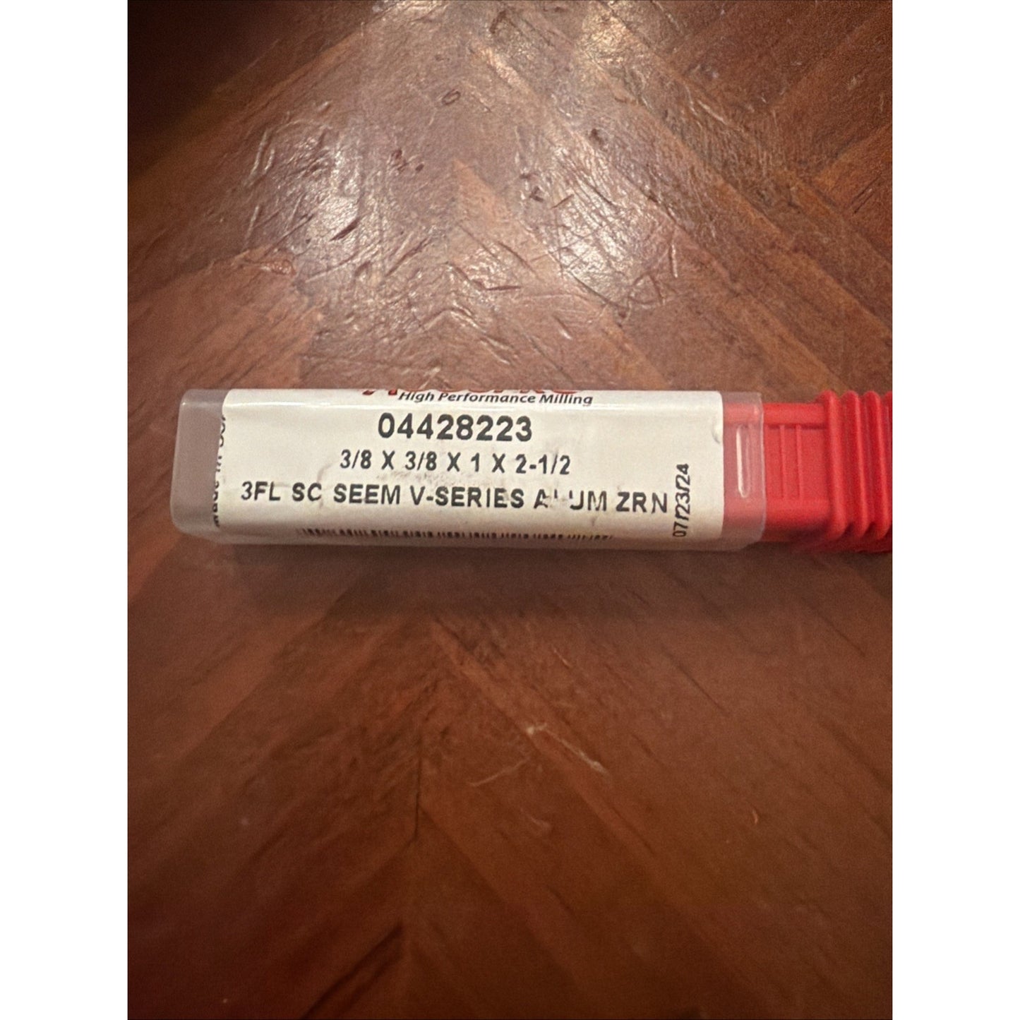 Clear plastic tool case with a white label displaying product number 04428223, dimensions 3/8 x 3/8 x 1 x 2-1/2, and text indicating a 3-flute solid carbide seam V-series aluminum zirconium milling cutter, resting on a wooden surface.