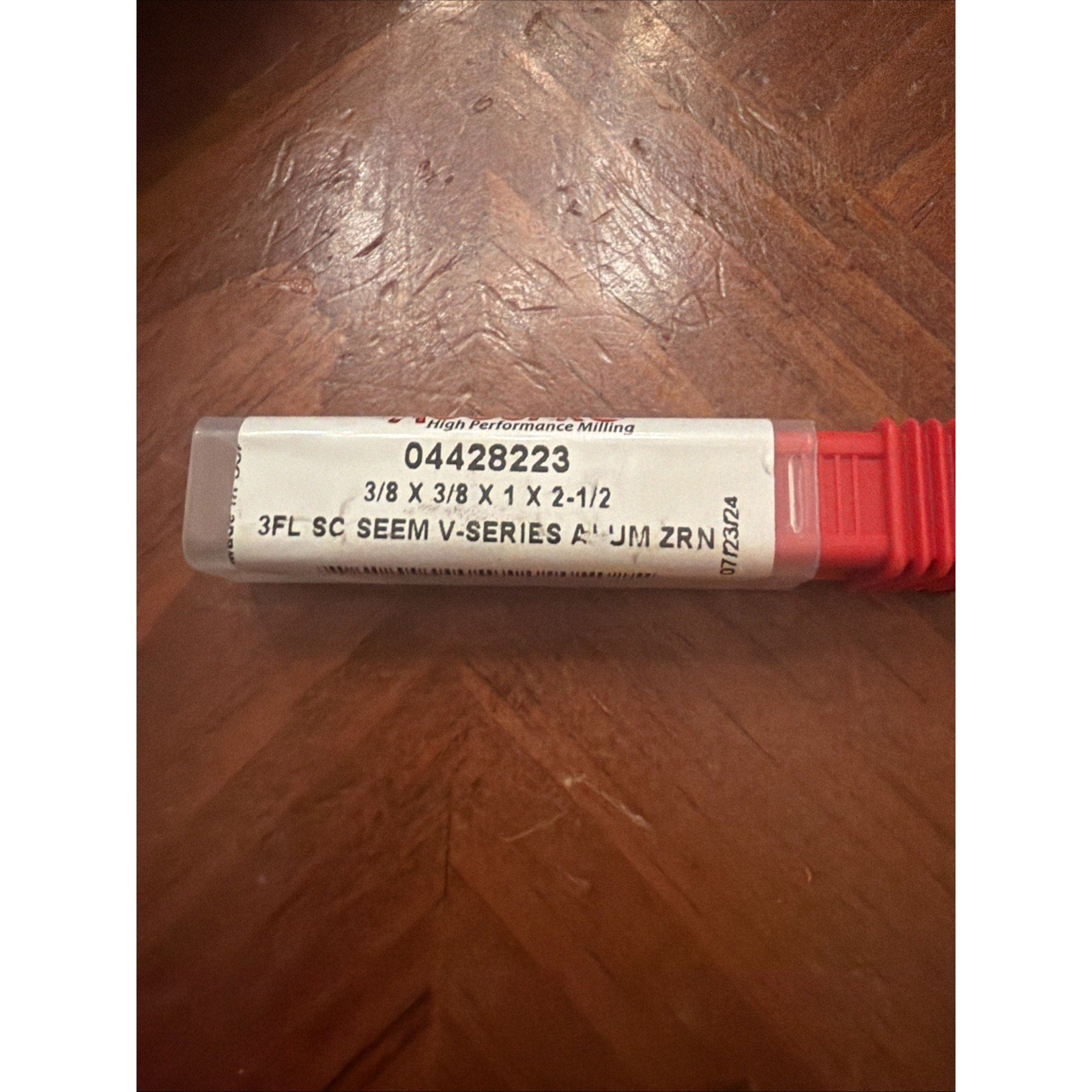 Clear plastic tool case with a white label displaying product number 04428223, dimensions 3/8 x 3/8 x 1 x 2-1/2, and text indicating a 3-flute solid carbide seam V-series aluminum zirconium milling cutter, resting on a wooden surface.