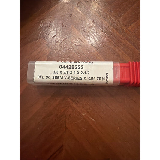 Clear plastic tool case with a white label displaying product number 04428223, dimensions 3/8 x 3/8 x 1 x 2-1/2, and text indicating a 3-flute solid carbide seam V-series aluminum zirconium milling cutter, resting on a wooden surface.