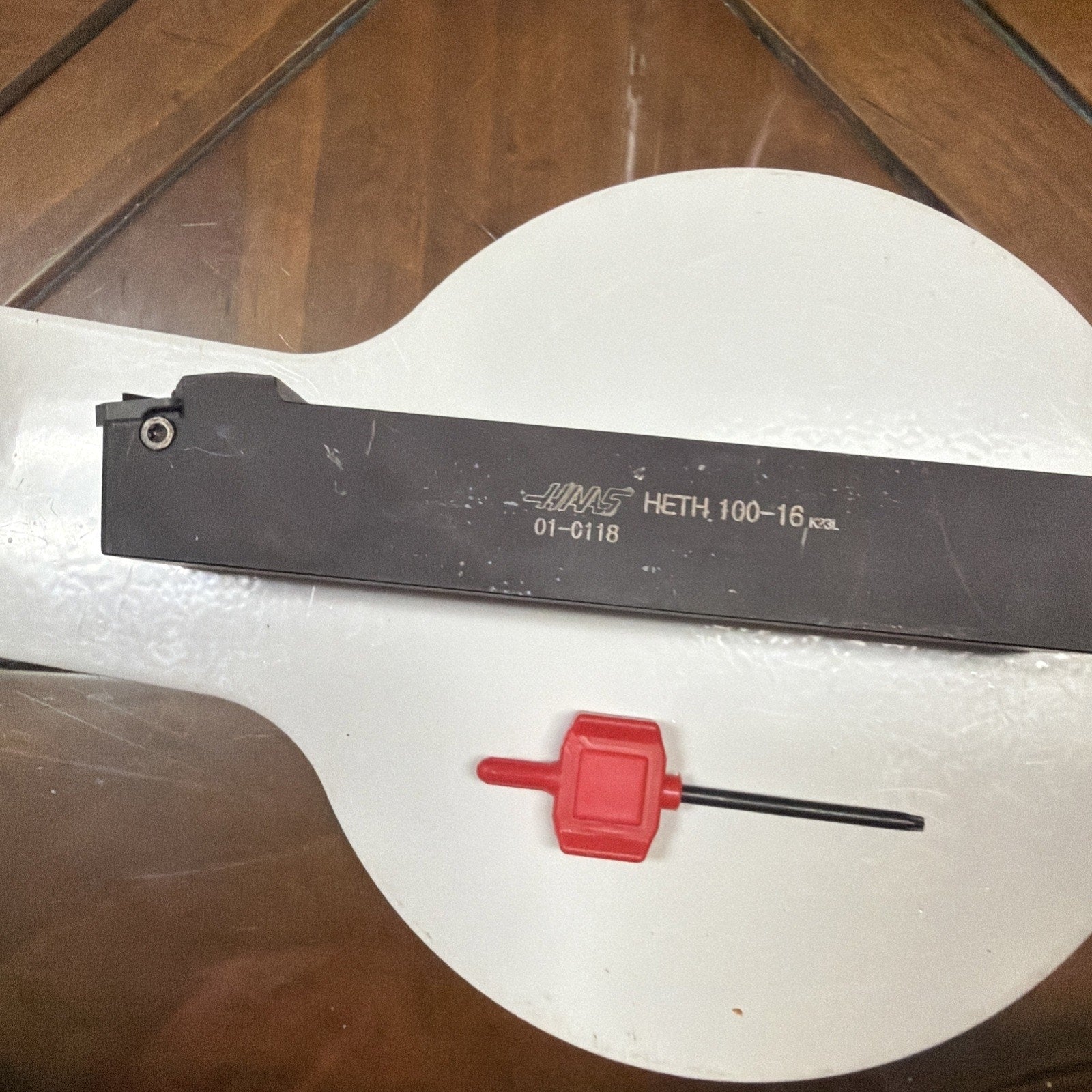 Rectangular black metal tool holder with engraved model numbers, paired with a small red-handled hex key, displayed on a white circular surface over a wooden background.