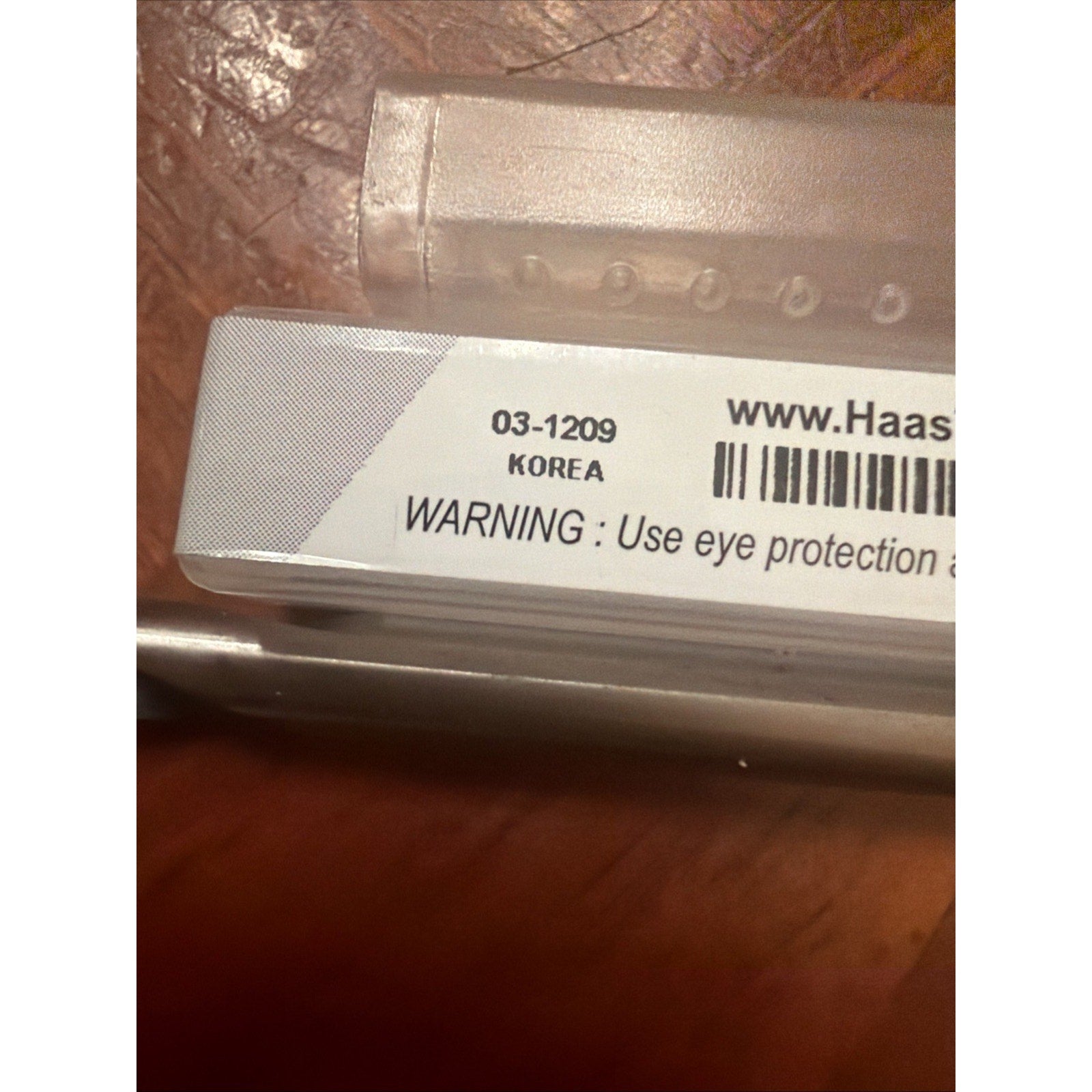 Close-up of a plastic container label showing product code 03-1209, country of origin Korea, part of a website URL, and a safety warning advising the use of eye protection.