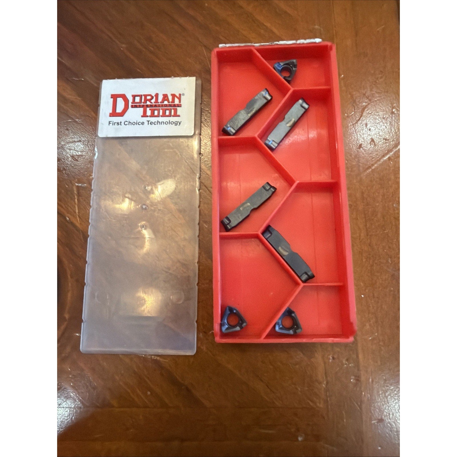 Red plastic compartmentalized tray holding six dark metal cutting inserts of varying shapes, next to a translucent plastic lid with a white label featuring red and black "Dorian Tool" branding, on a wooden surface.
