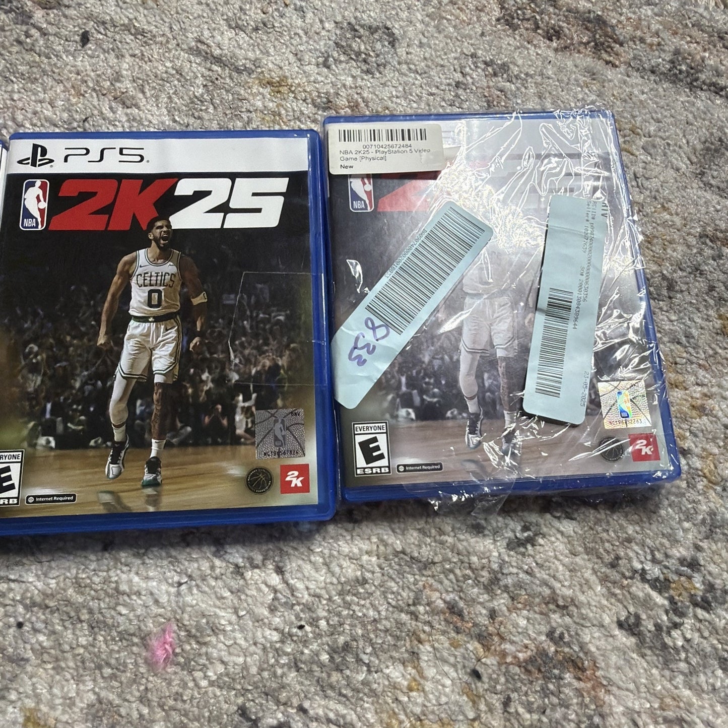 Two PlayStation 5 NBA 2K25 game cases on a carpeted floor, one showing the front cover with a basketball player in a Celtics uniform, the other showing the back with multiple barcode stickers and plastic wrapping.