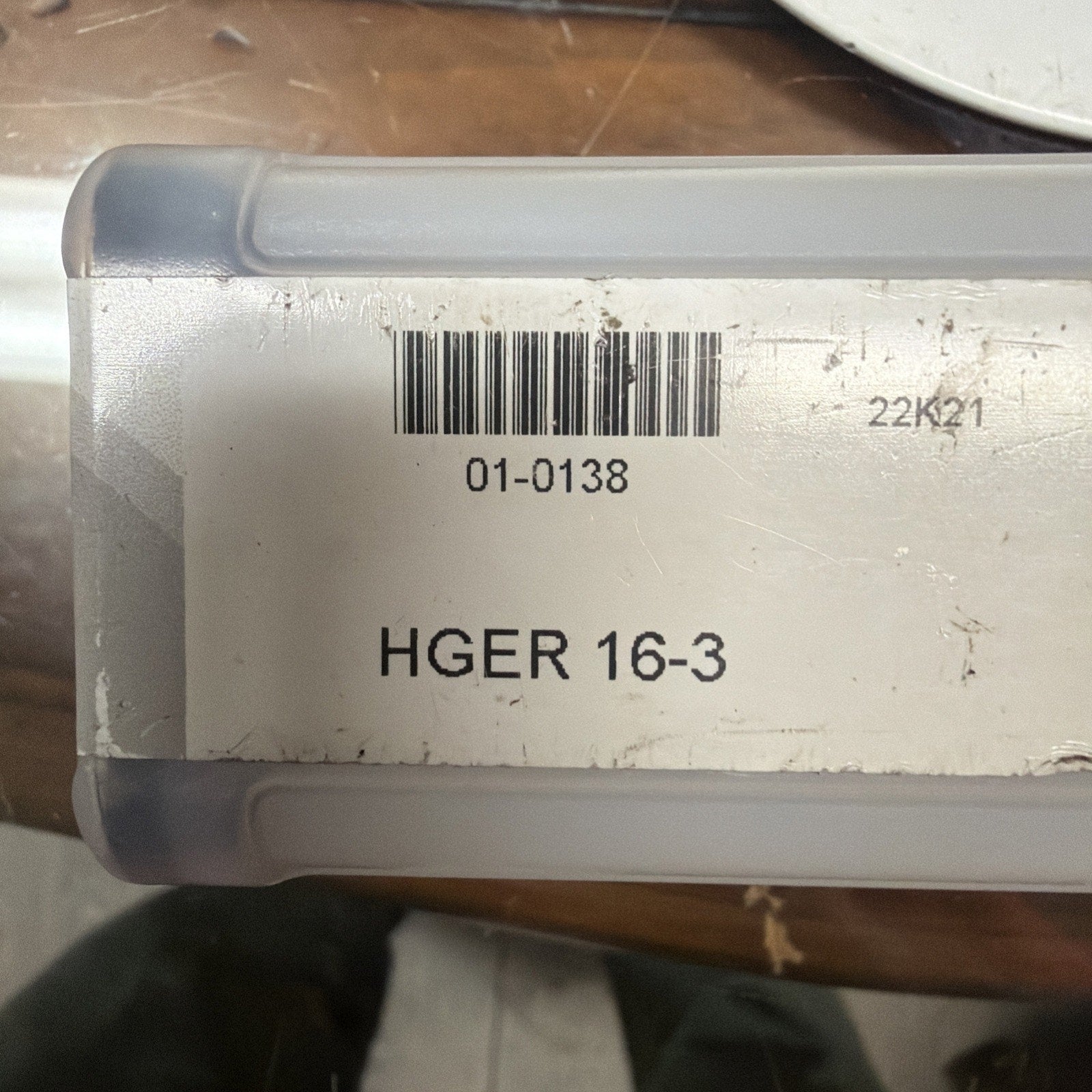 Close-up of a white and gray rectangular plastic container with a barcode, product code "01-0138," and printed text "HGER 16-3" on a slightly worn label, resting on a wooden surface.