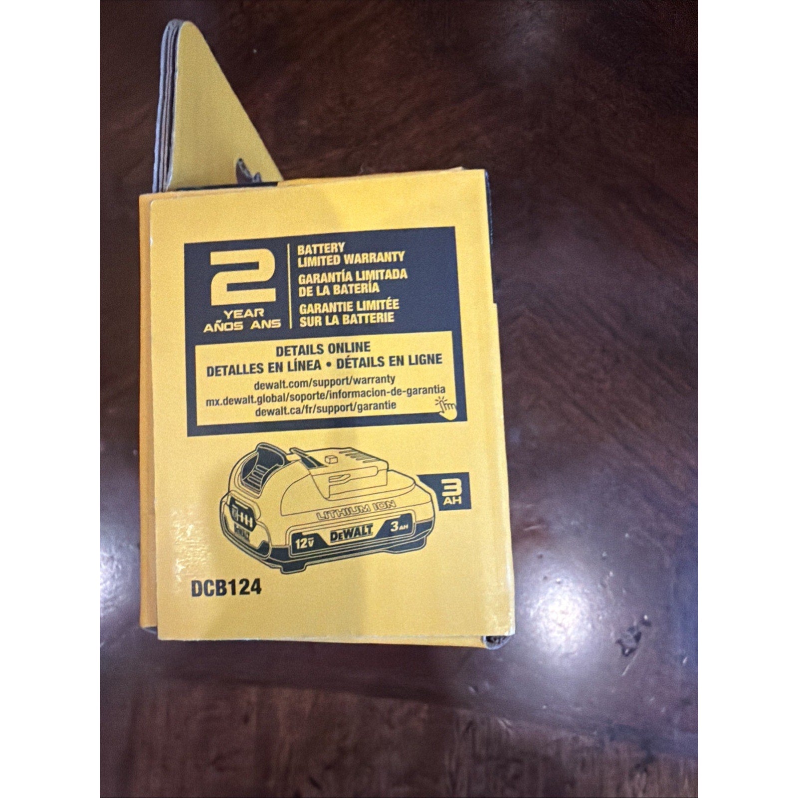 Yellow product box featuring an illustration of a 12V 3Ah DeWalt battery, warranty information in English, Spanish, and French, and a model number DCB124 printed on a dark wooden surface.