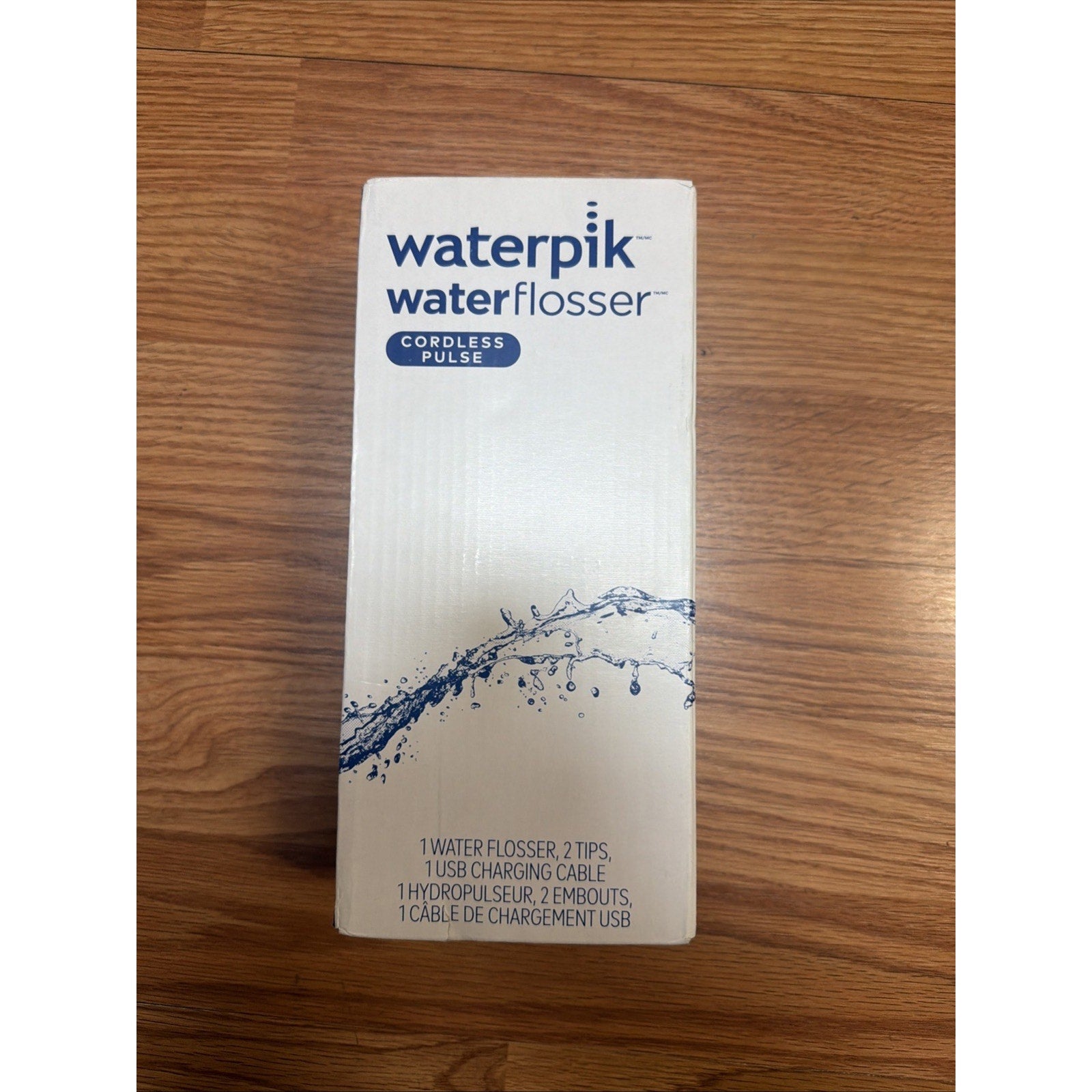 White rectangular box packaging for a Waterpik Water Flosser Cordless Pulse with blue branding and a splash of water graphic on a wooden surface.