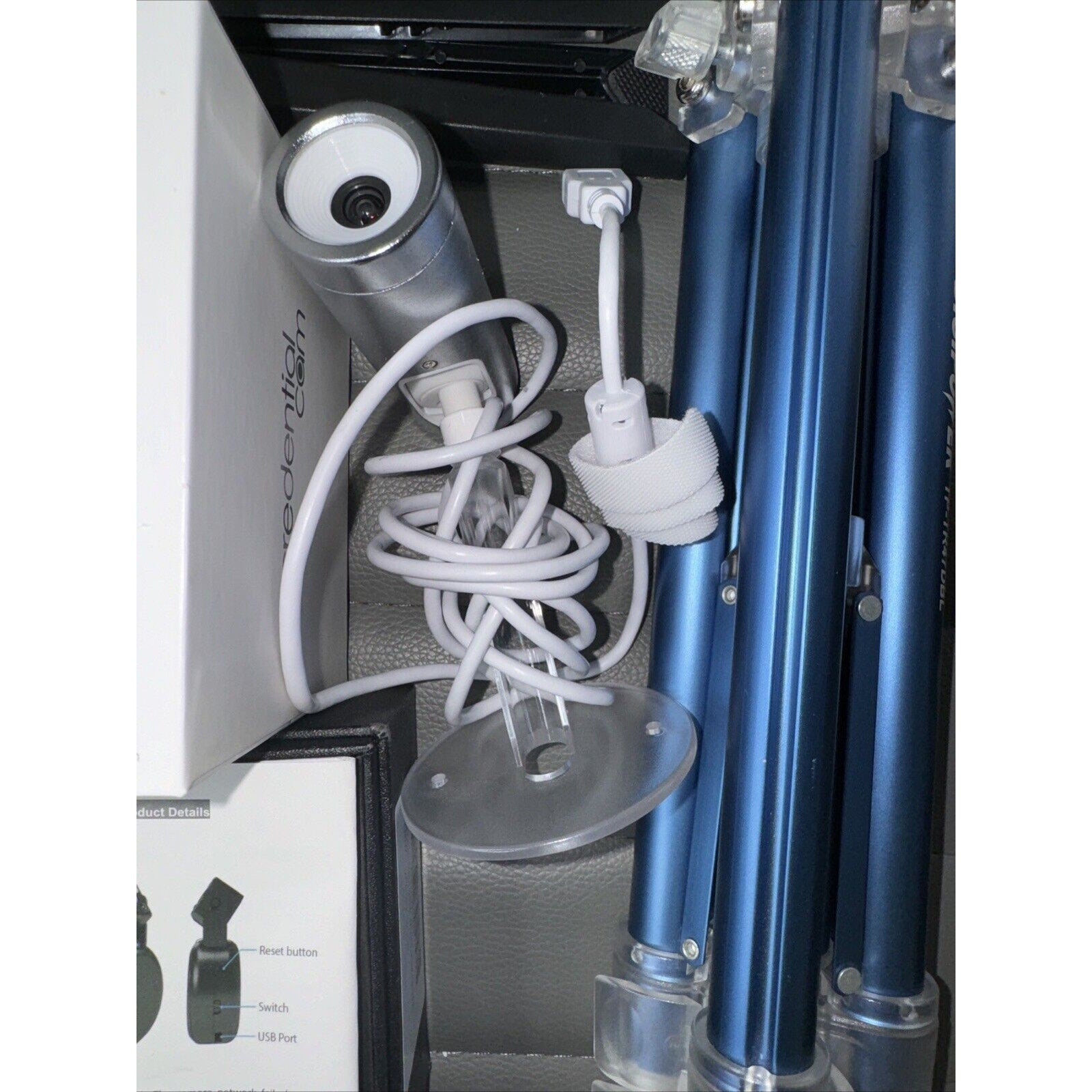 Lot of photography equipment including silver camera, coiled USB cable, blue tripods, and white packaging boxes arranged in storage case