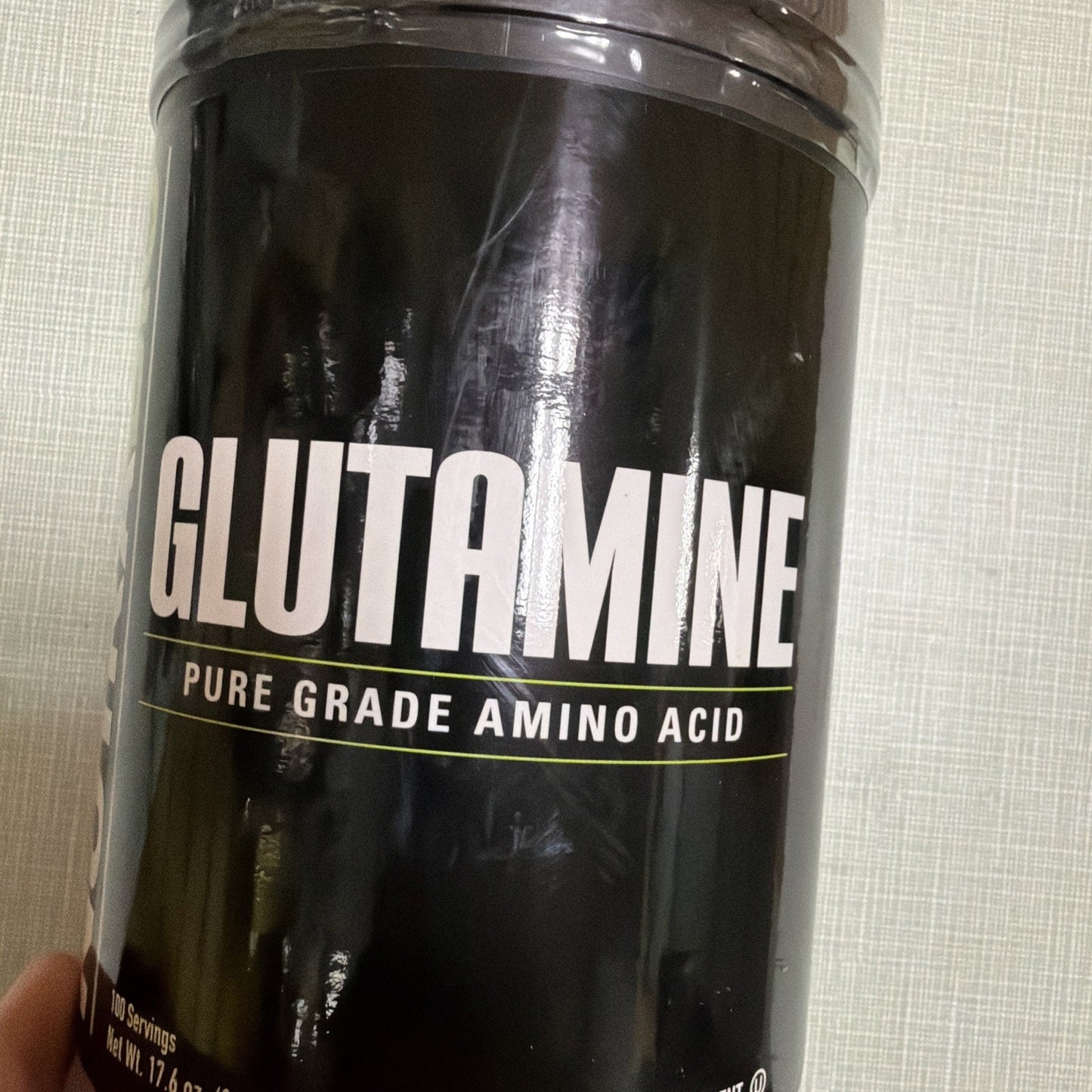 Black container labeled "Glutamine" with text indicating a pure grade amino acid supplement, held against a neutral textured background.