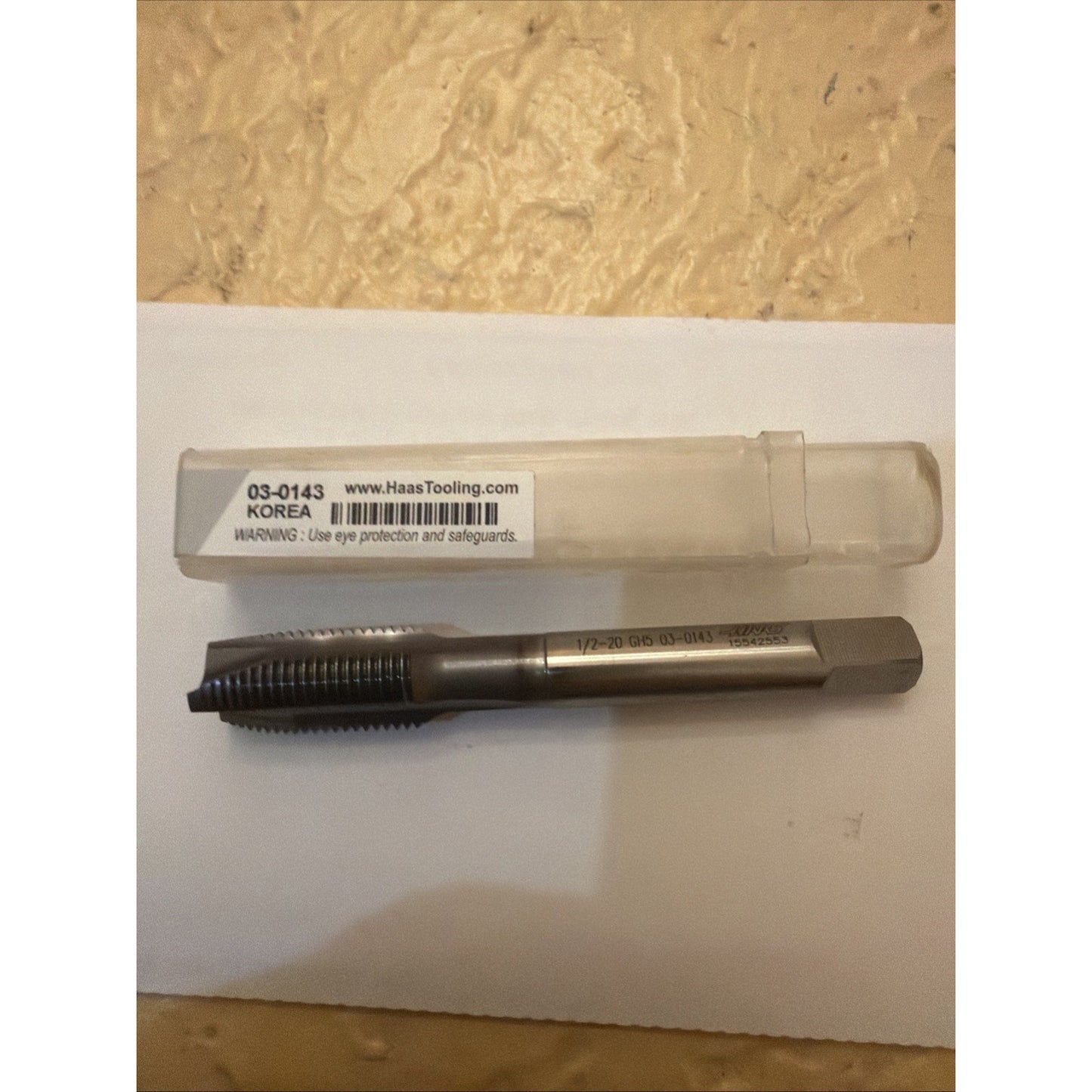 Steel threading tap with sharp cutting threads and a hexagonal shank, placed on a white sheet next to a clear plastic storage case labeled with product code and safety warning.