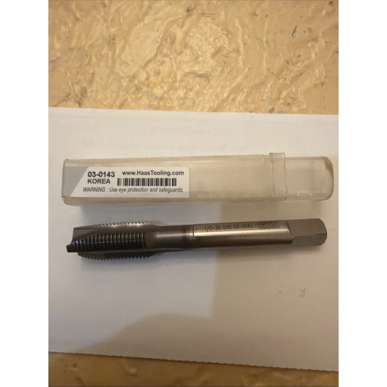 Steel threading tap with sharp cutting threads and a hexagonal shank, placed on a white sheet next to a clear plastic storage case labeled with product code and safety warning.