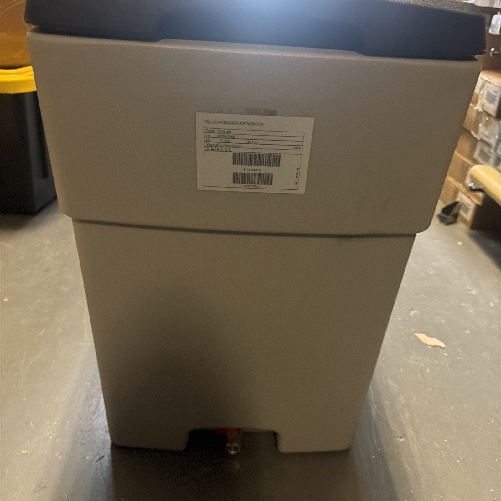 Gray rectangular container with a black lid and a white label on the front, placed on a concrete floor in a storage or industrial setting.