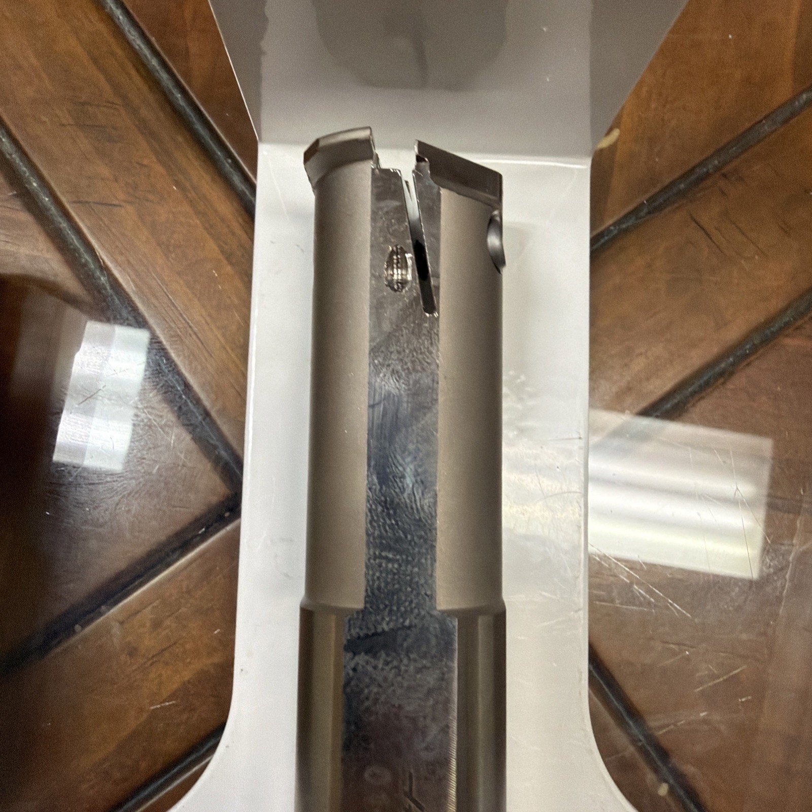 Metallic cylindrical object with a notch and a small screw on its surface, placed inside a white open box on a wooden table with a glass top reflecting light.