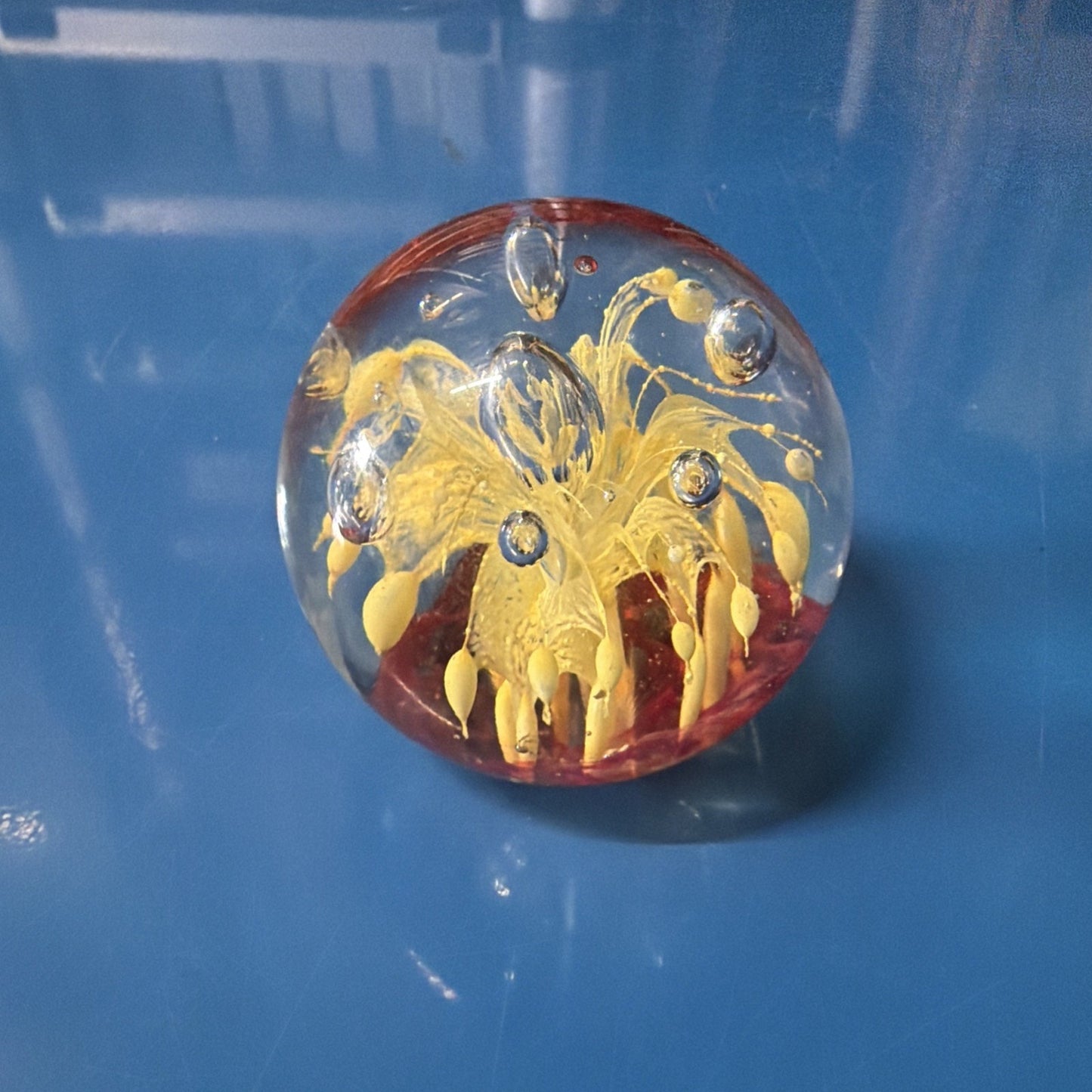 Clear glass paperweight with intricate yellow flower-like design and bubbles suspended inside, resting on a smooth blue surface.