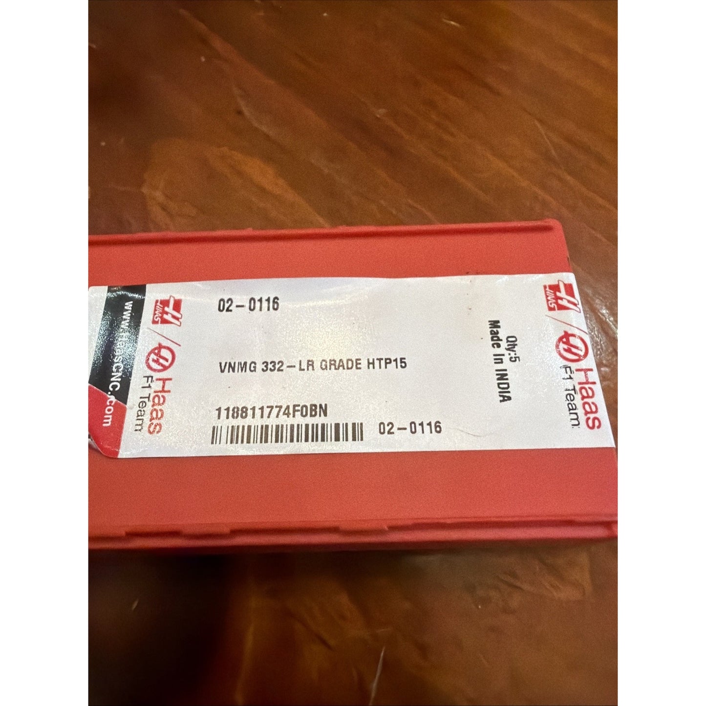 Red plastic storage case with a white label displaying product code "VNMG 332-LR GRADE HTP15," barcode, manufacturing origin "Made in India," and Haas F1 Team branding, placed on a wooden surface.