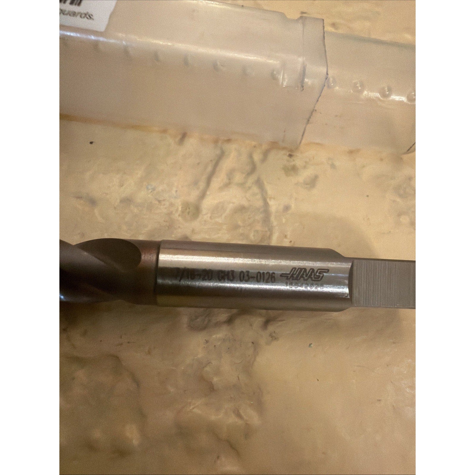 Close-up of a metal threading tap with engraved product codes and branding on its smooth shank, resting on a textured beige surface near a translucent plastic storage tube.