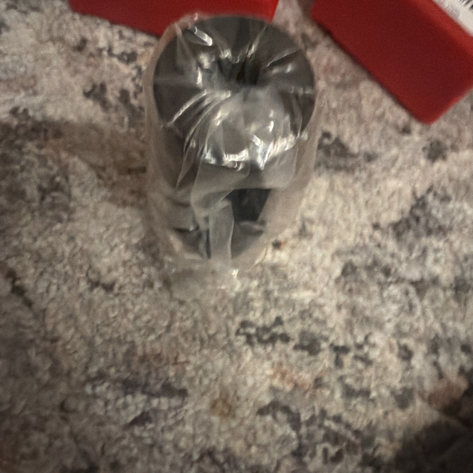 Small black cylindrical metal component wrapped in clear plastic, positioned upright on a textured gray and beige carpet surface with red plastic containers nearby.
