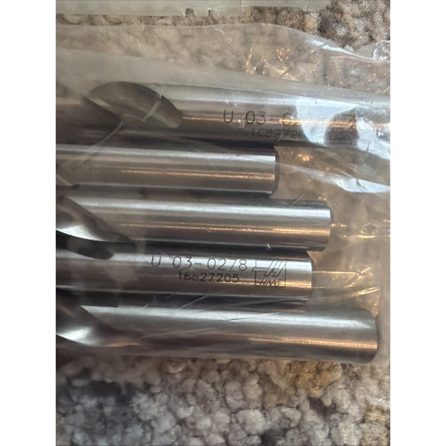 Several smooth silver cylindrical metal rods with engraved numerical markings, partially contained in a clear plastic bag, resting on a textured light-colored fabric surface.