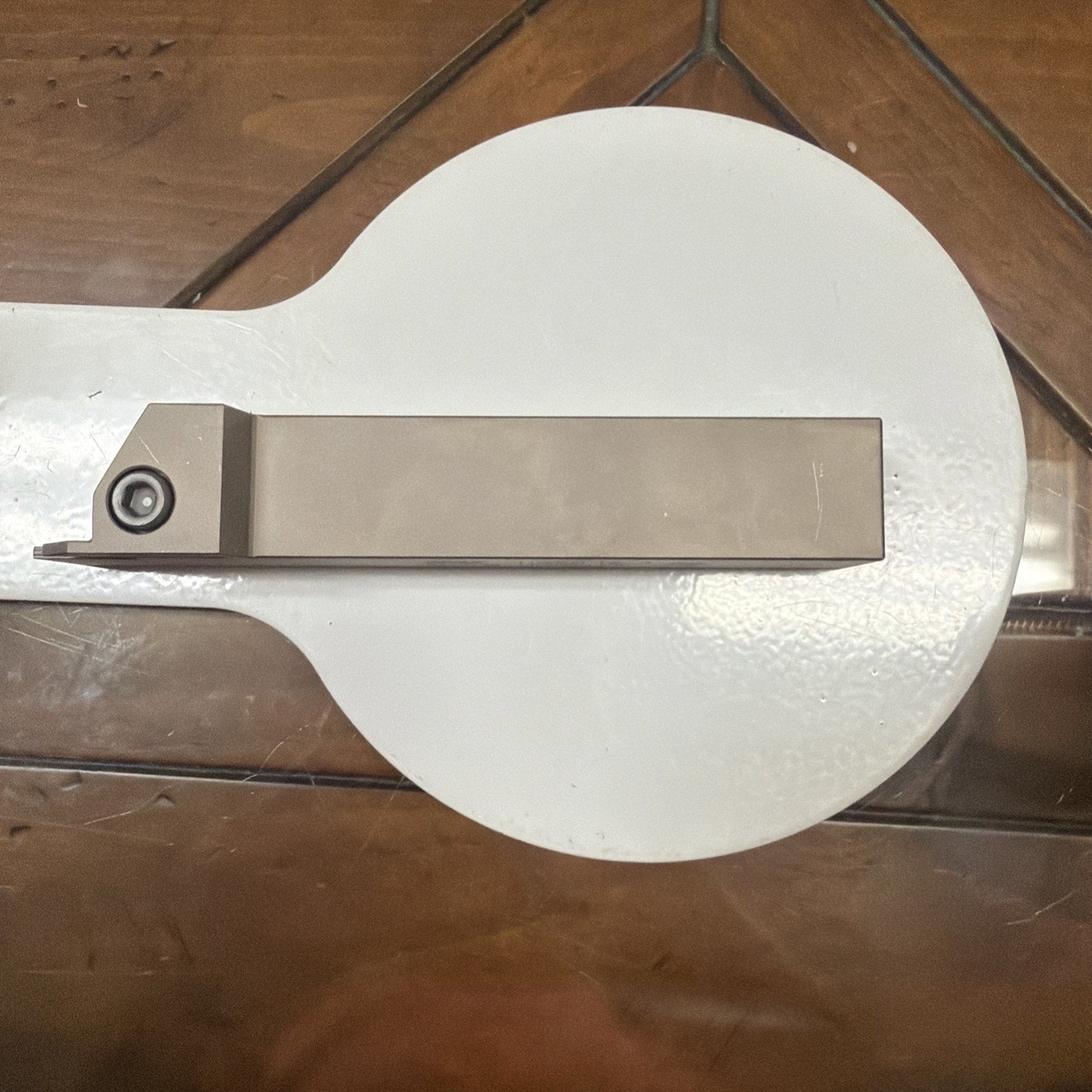 Rectangular metal cutting tool with a carbide insert secured by a screw, resting horizontally on a round white surface atop a wooden table.