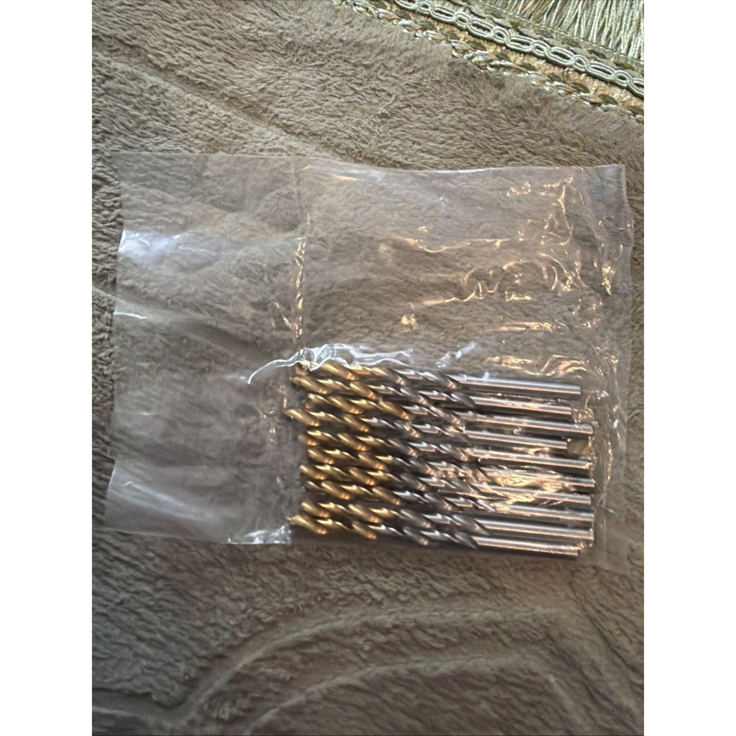 Set of ten metallic drill bits with spiral flutes and shiny silver shanks, arranged side by side inside a partially open clear plastic bag on a textured fabric surface.