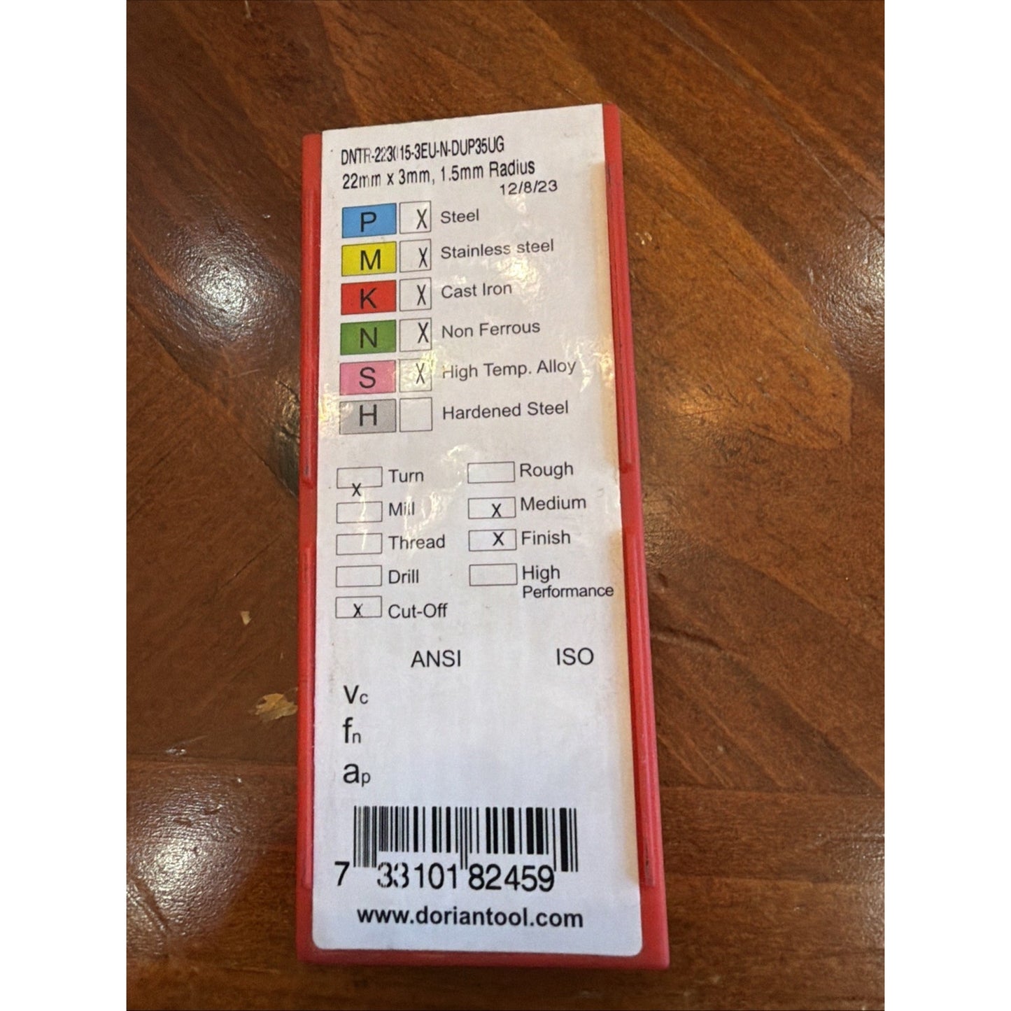 Rectangular red plastic case with a white label displaying color-coded metal type classifications, machining operation checkboxes, ANSI and ISO markings, and a barcode on a wooden surface.