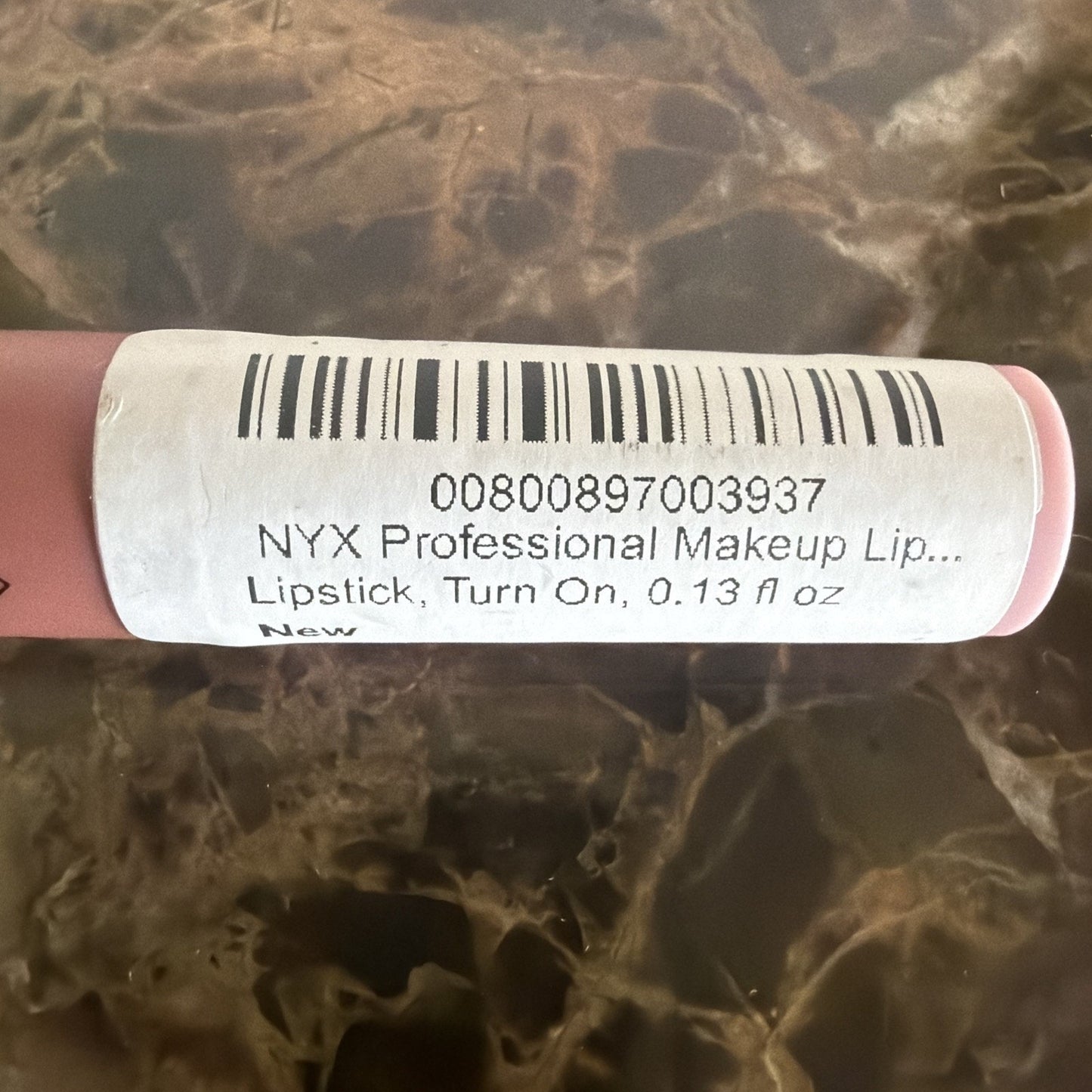 Close-up view of a pink NYX Professional Makeup lipstick tube with a white barcode label showing product details on a marble surface background.