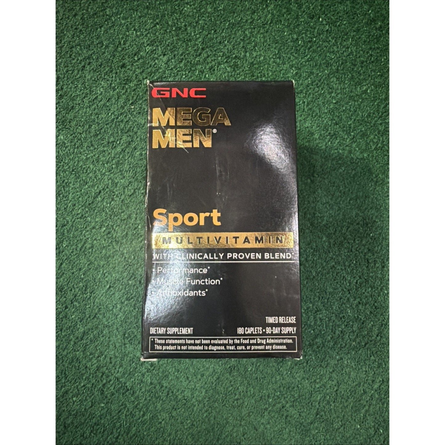 Black rectangular box of GNC Mega Men Sport multivitamin dietary supplement with gold and white text highlighting performance, muscle function, and antioxidants, against a green textured background.