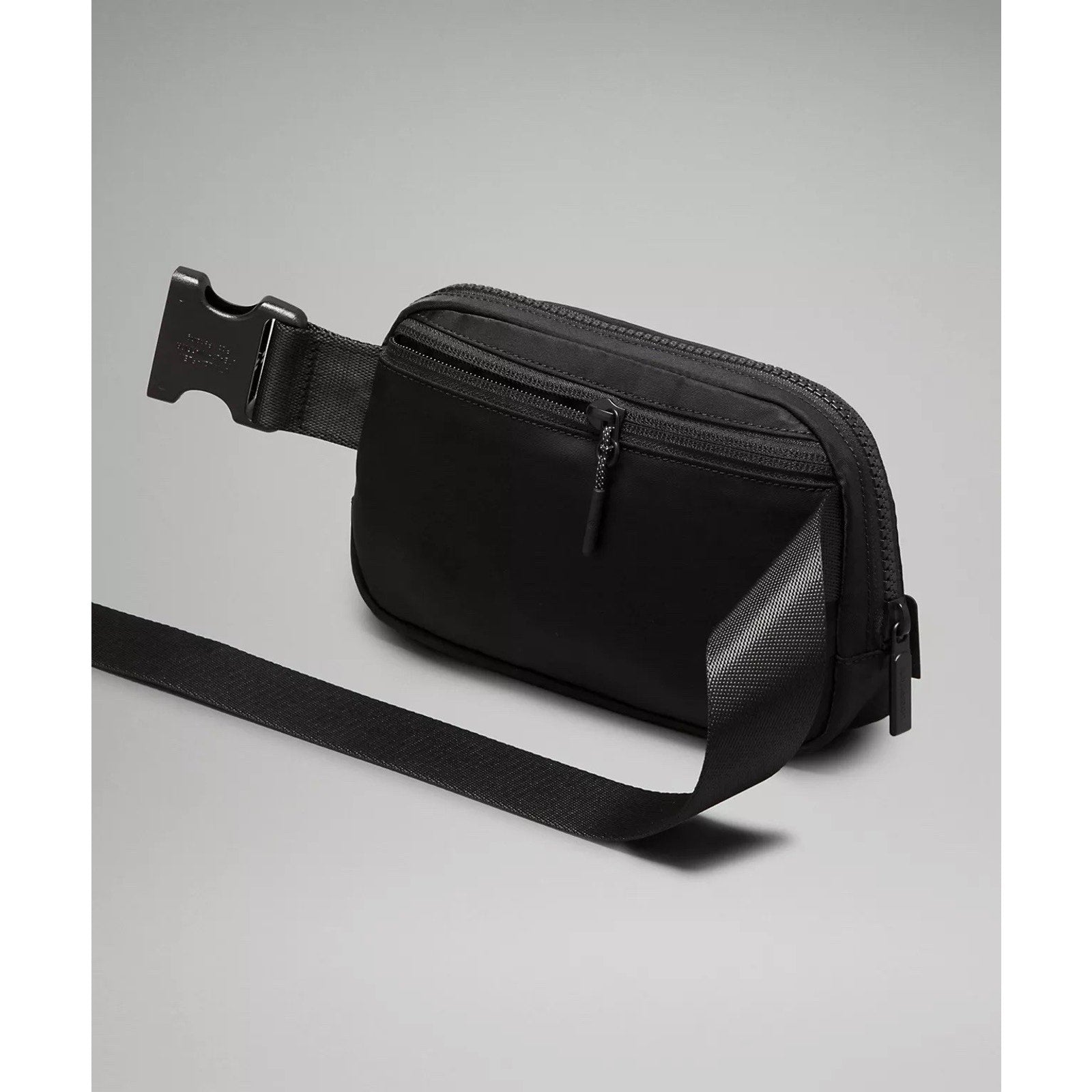 Black compact waist bag with front zip pocket, adjustable strap with plastic buckle, and smooth matte finish on a plain gray background.