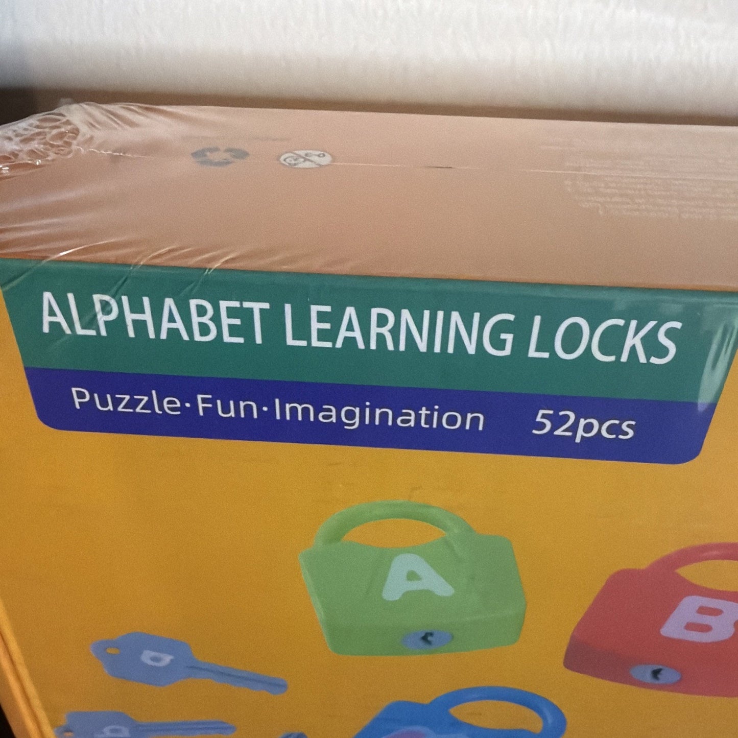 Corner of yellow toy box packaging displaying "Alphabet Learning Locks" with colorful lock and key illustrations along with text highlighting puzzle, fun, and imagination features plus 52 pieces included.