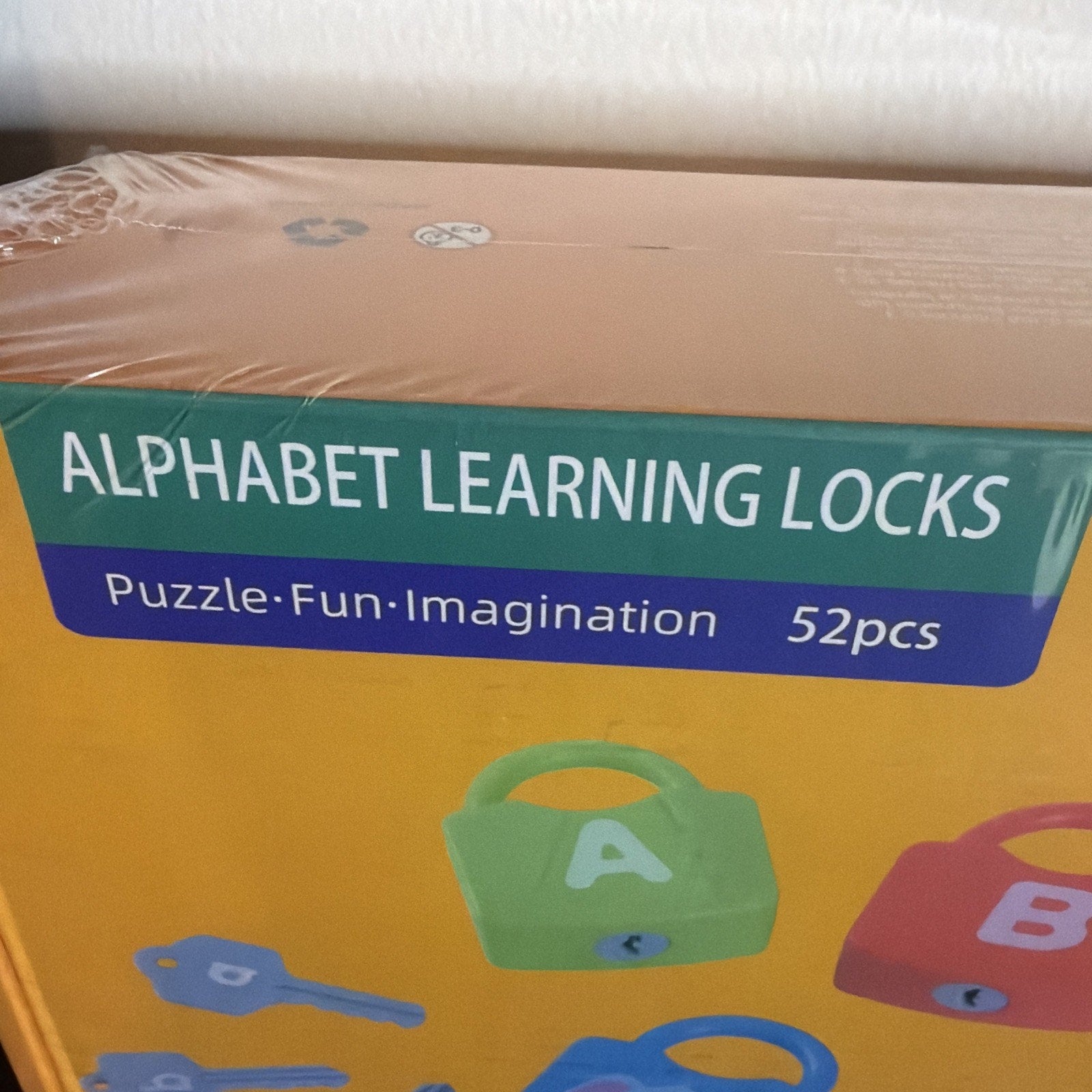 Corner of yellow toy box packaging displaying "Alphabet Learning Locks" with colorful lock and key illustrations along with text highlighting puzzle, fun, and imagination features plus 52 pieces included.