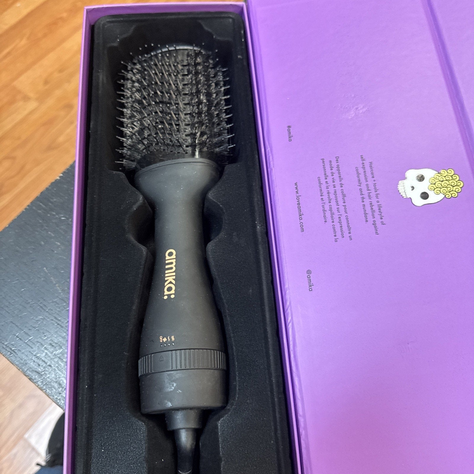Amika Hair Blow Dryer Brush in black with gold logo, displayed in purple branded packaging box