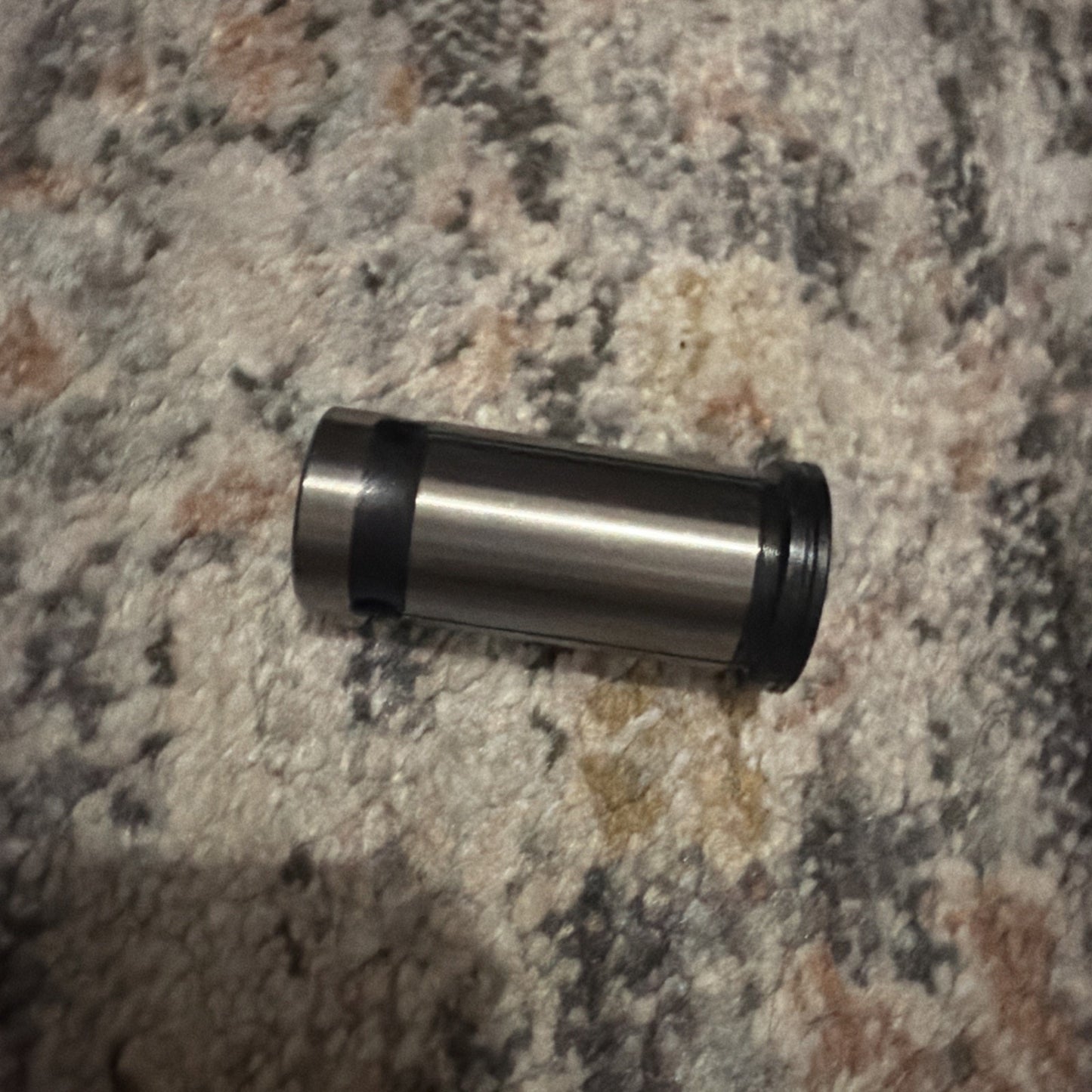 Metal cylindrical tool component with a smooth surface, featuring a black ring near one end, resting on a multicolored textured carpet background.