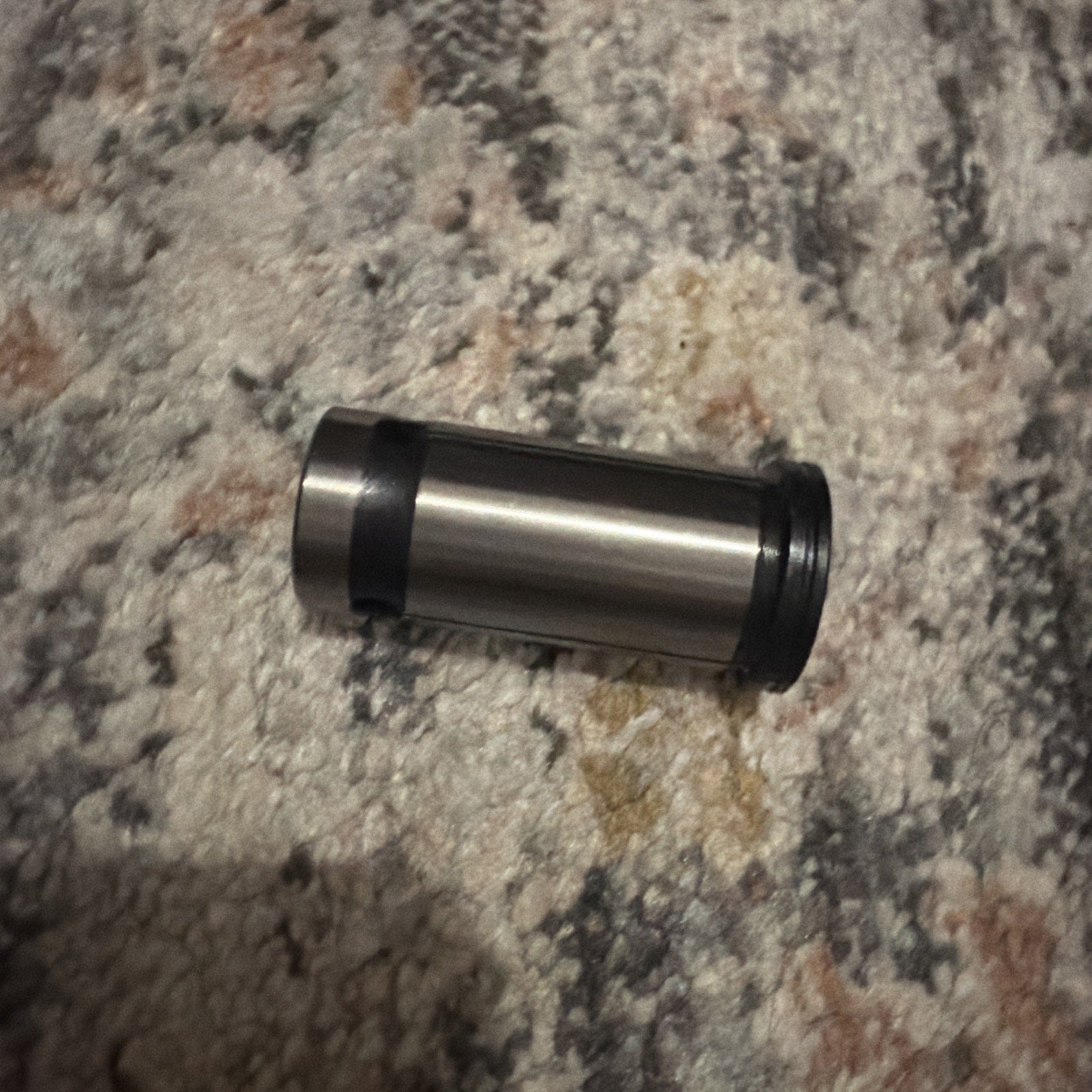 Metal cylindrical tool component with a smooth surface, featuring a black ring near one end, resting on a multicolored textured carpet background.