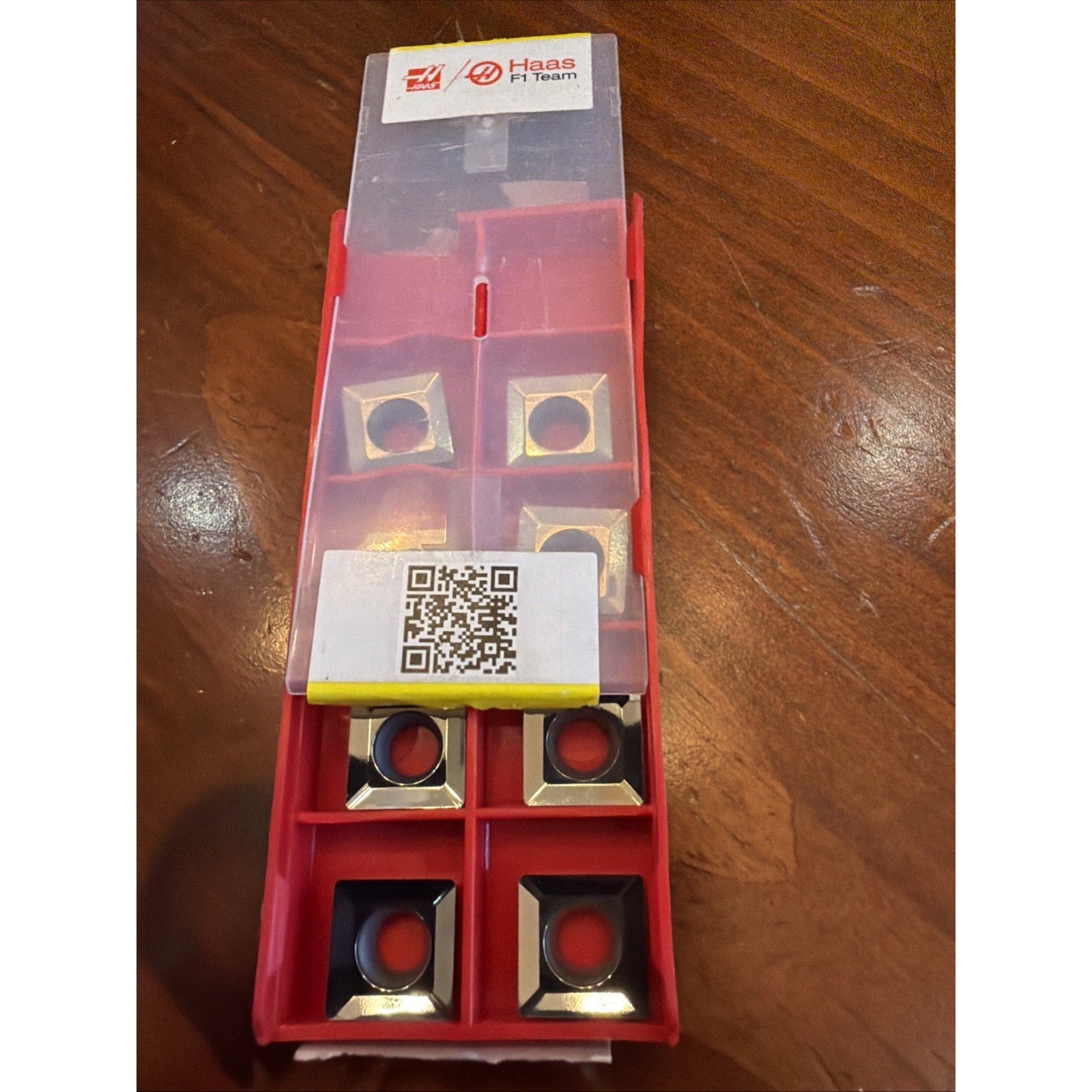 Red plastic storage box with a translucent lid partially open, containing eight square metal cutting inserts with central holes, placed on a wooden surface, featuring Haas F1 Team branding and a QR code label.