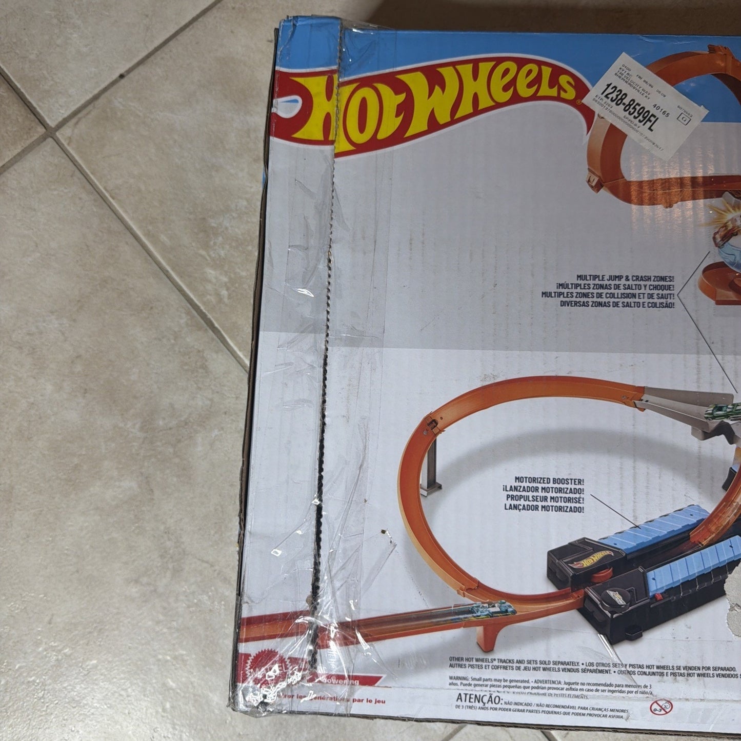 Partial view of a Hot Wheels toy car track packaging showing an orange looped racetrack with a motorized booster and launch pad, placed on a tiled floor.