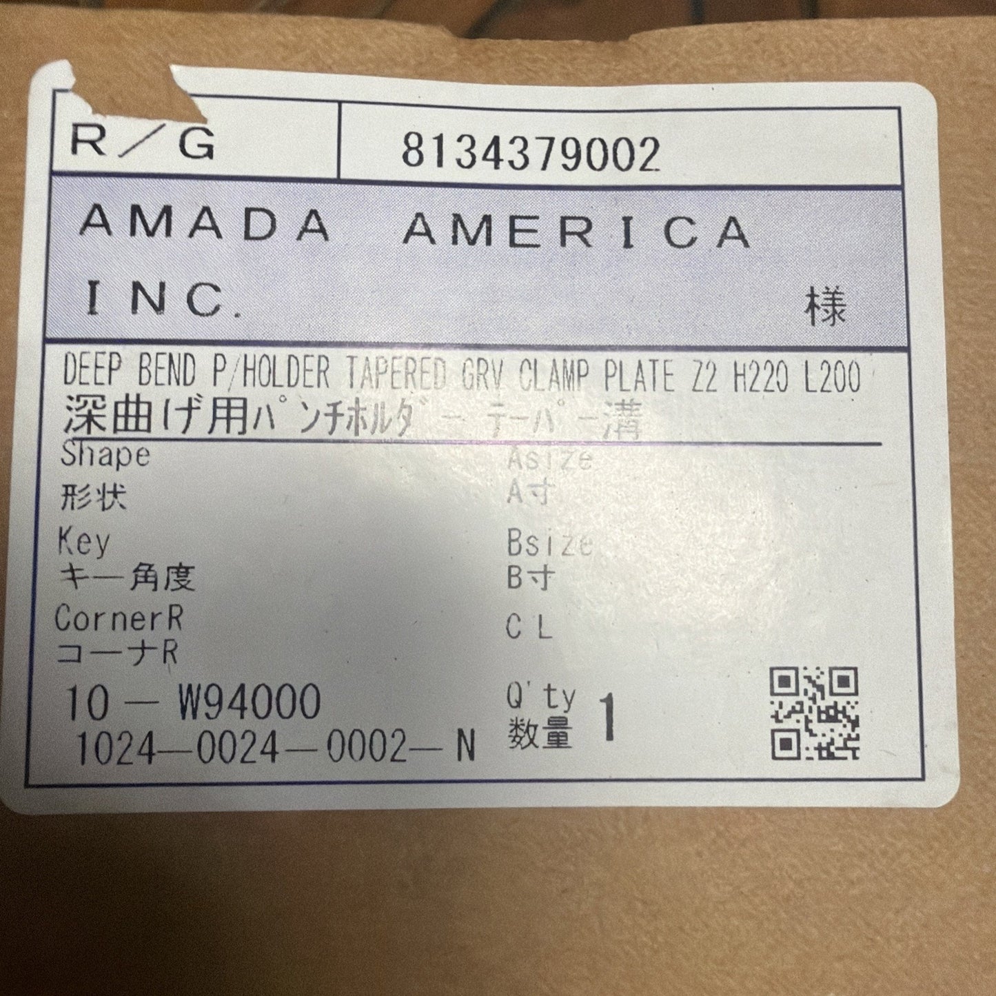 Close-up of a white label on brown cardboard displaying printed product information including a company name, part description, specifications, quantity, and a QR code.