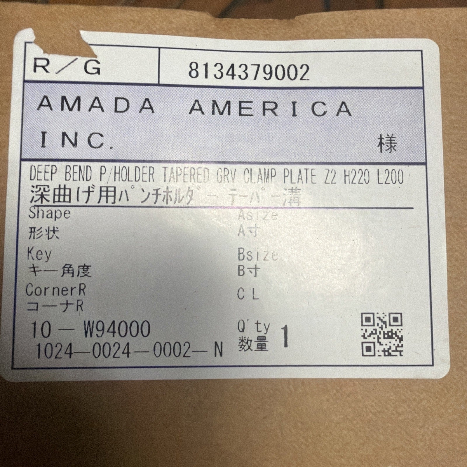 Close-up of a white label on brown cardboard displaying printed product information including a company name, part description, specifications, quantity, and a QR code.