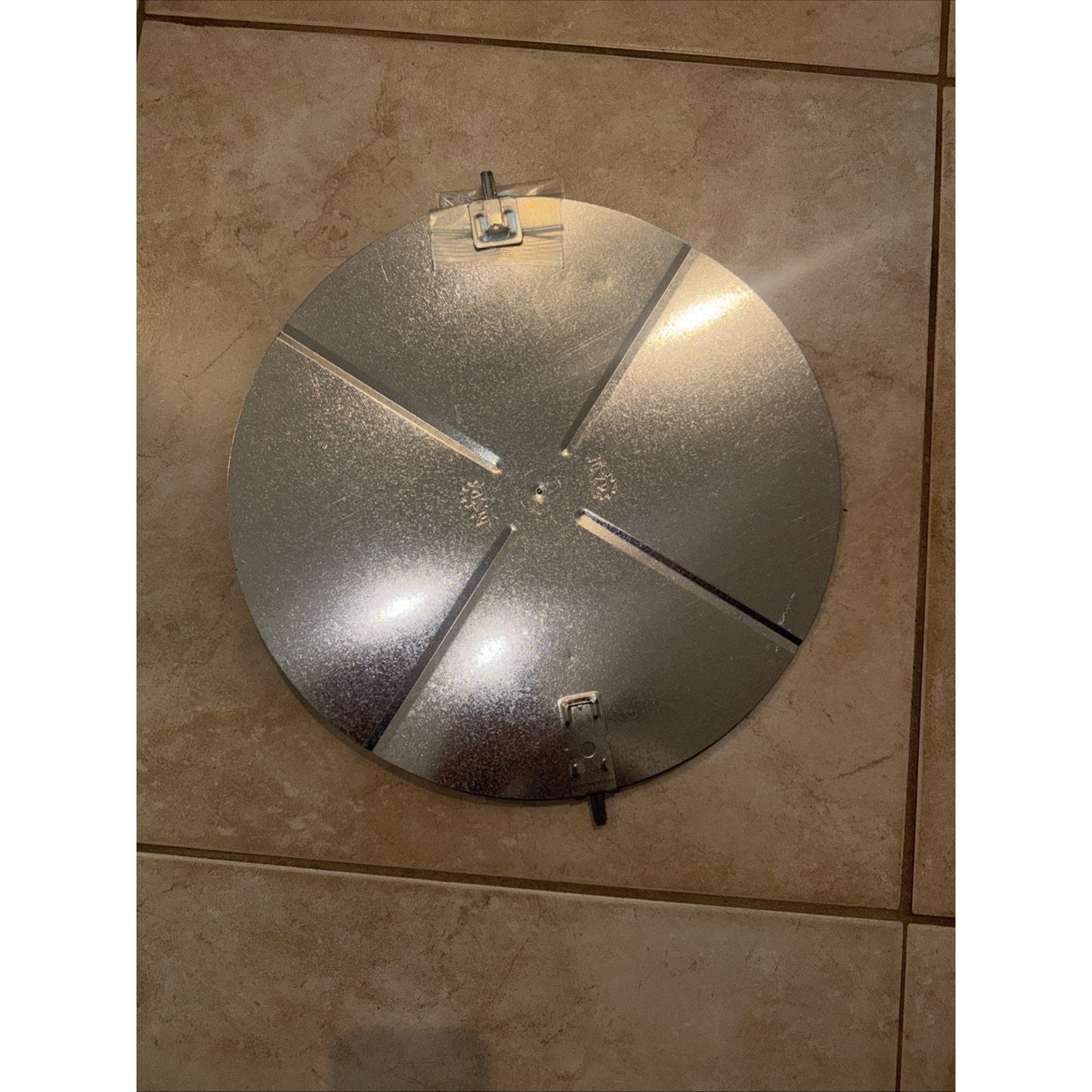 Round metallic silver reflector with four equally spaced cutouts and three metal clips around the edge, placed on a beige tiled floor.