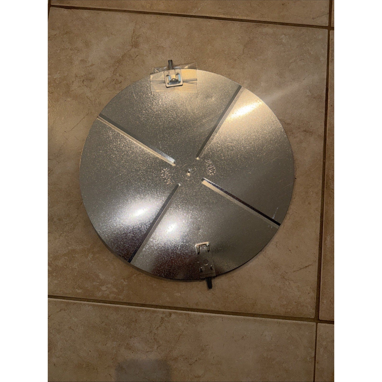 Round metallic silver reflector with four equally spaced cutouts and three metal clips around the edge, placed on a beige tiled floor.