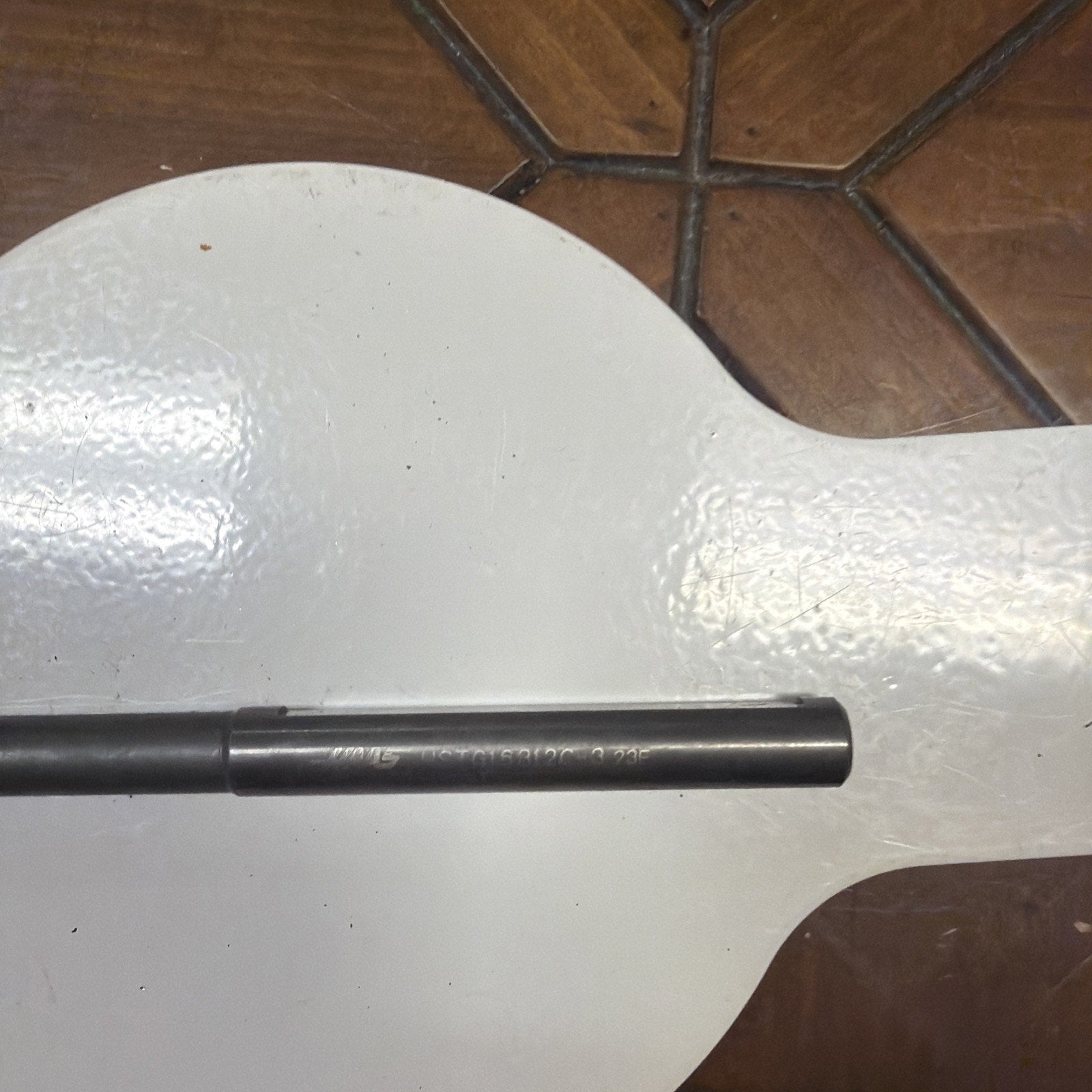 Close-up of a cylindrical metal rod with engraved alphanumeric markings, resting horizontally on a glossy white surface with brown tiled flooring in the background.