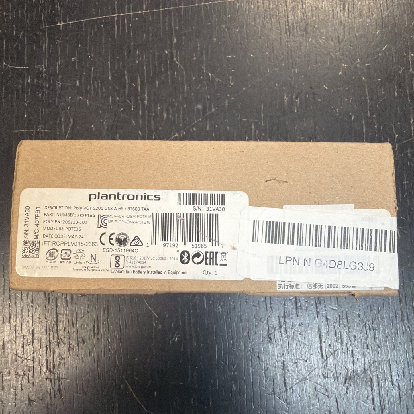 Brown rectangular cardboard box with a white printed label displaying product details, barcodes, and certification symbols, placed on a dark textured surface.