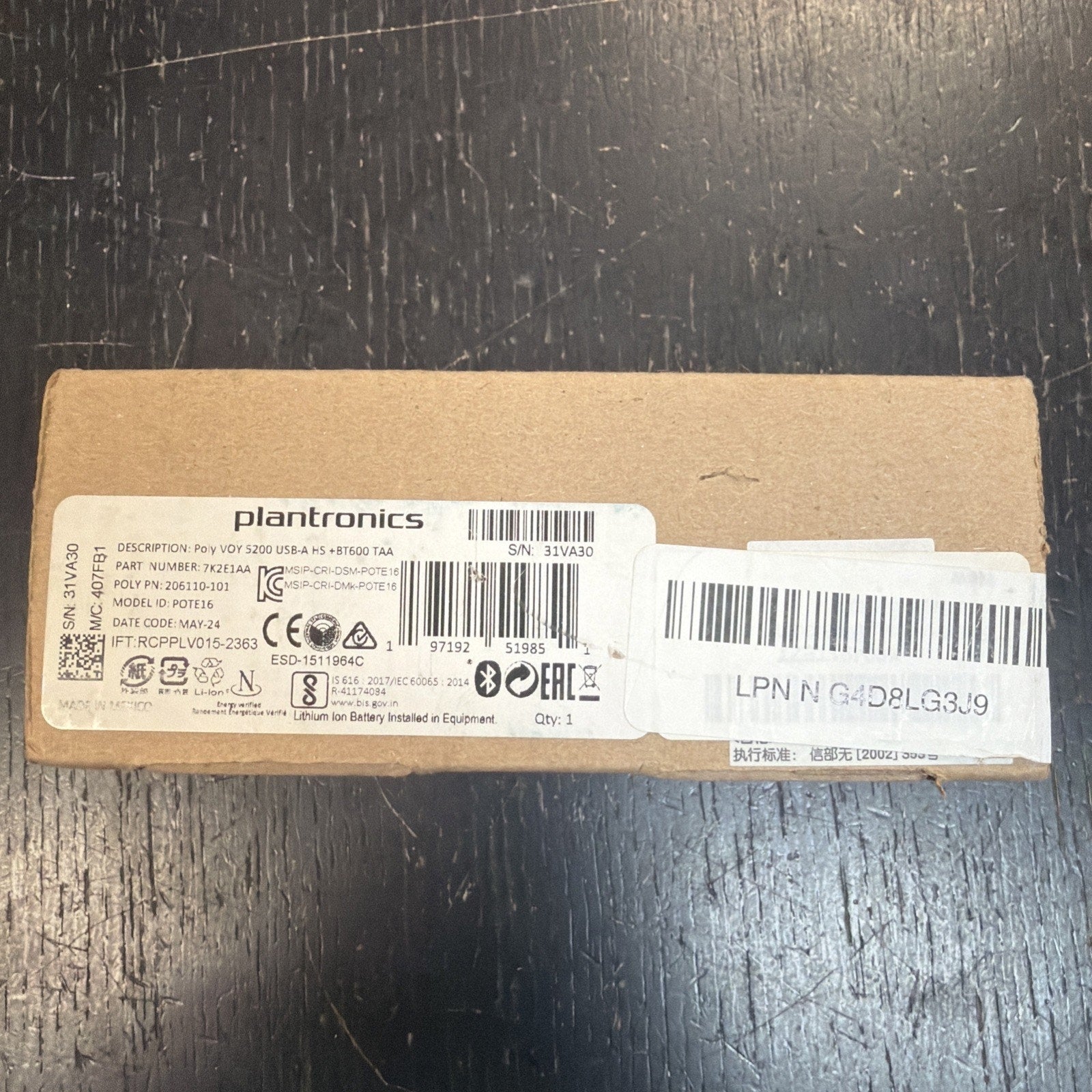 Brown rectangular cardboard box with a white printed label displaying product details, barcodes, and certification symbols, placed on a dark textured surface.