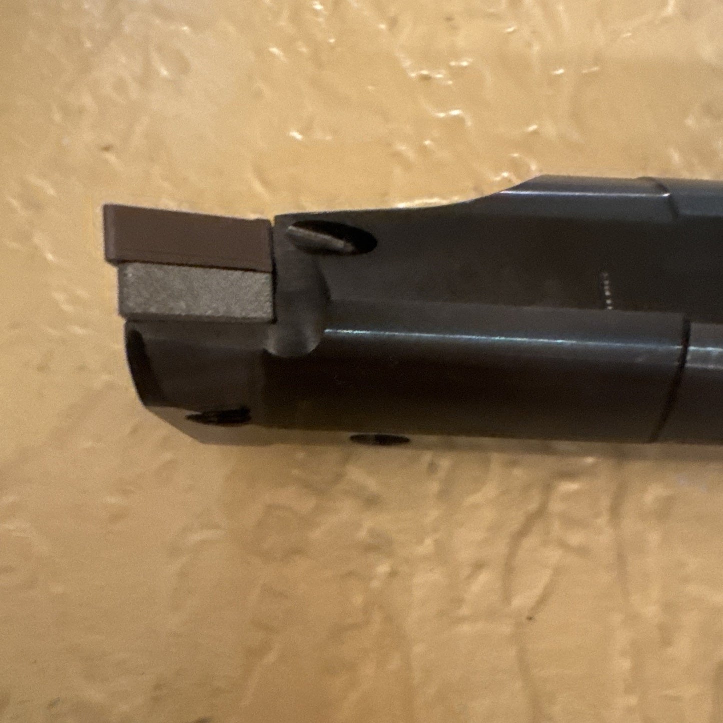 Close-up view of a dark metal cutting tool tip featuring a rectangular insert with a flat edge, positioned against a textured beige wall background.