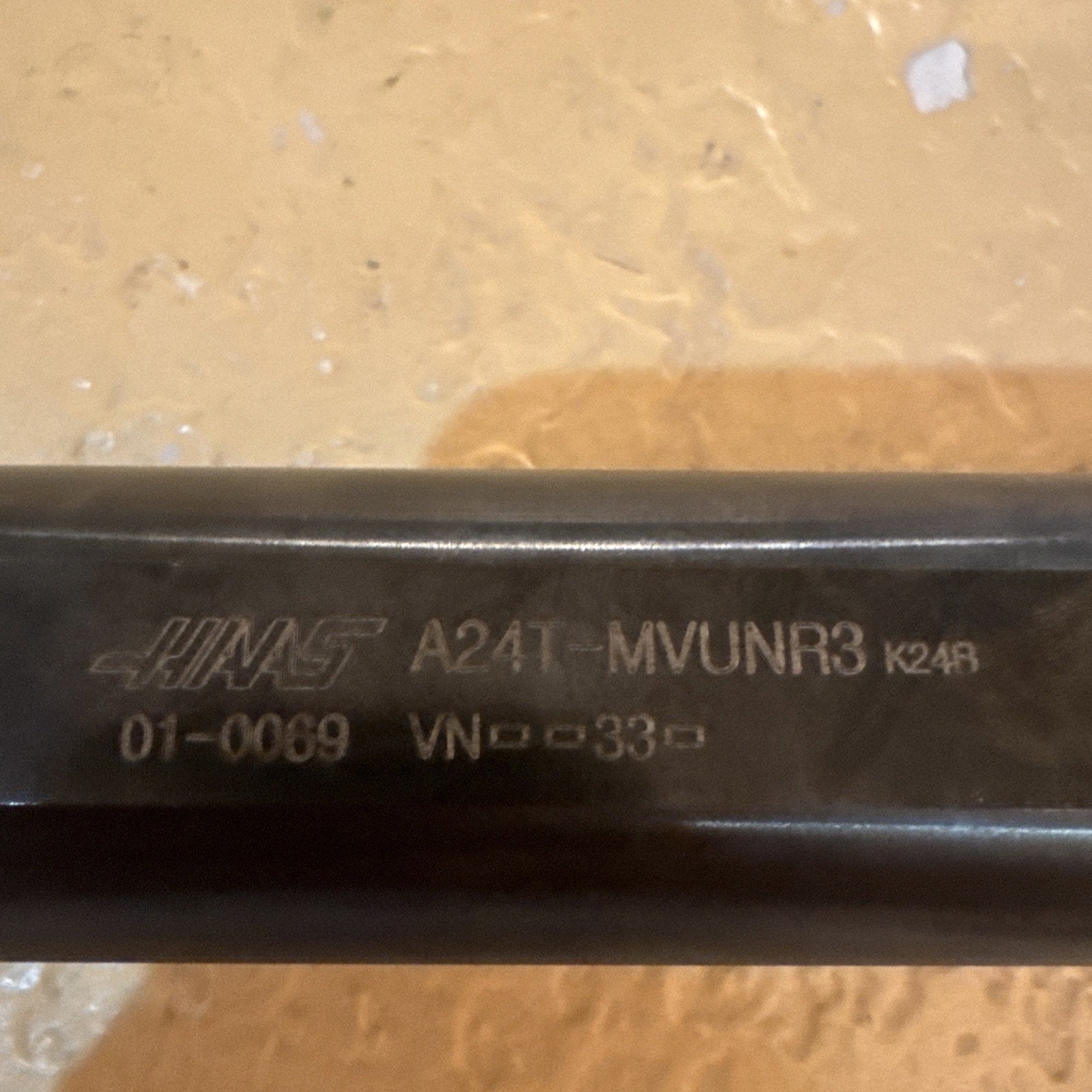 Close-up of a dark metal tool shaft engraved with the Haas logo, model number A24T-MVUNR3, and additional numeric codes on a textured light brown surface.