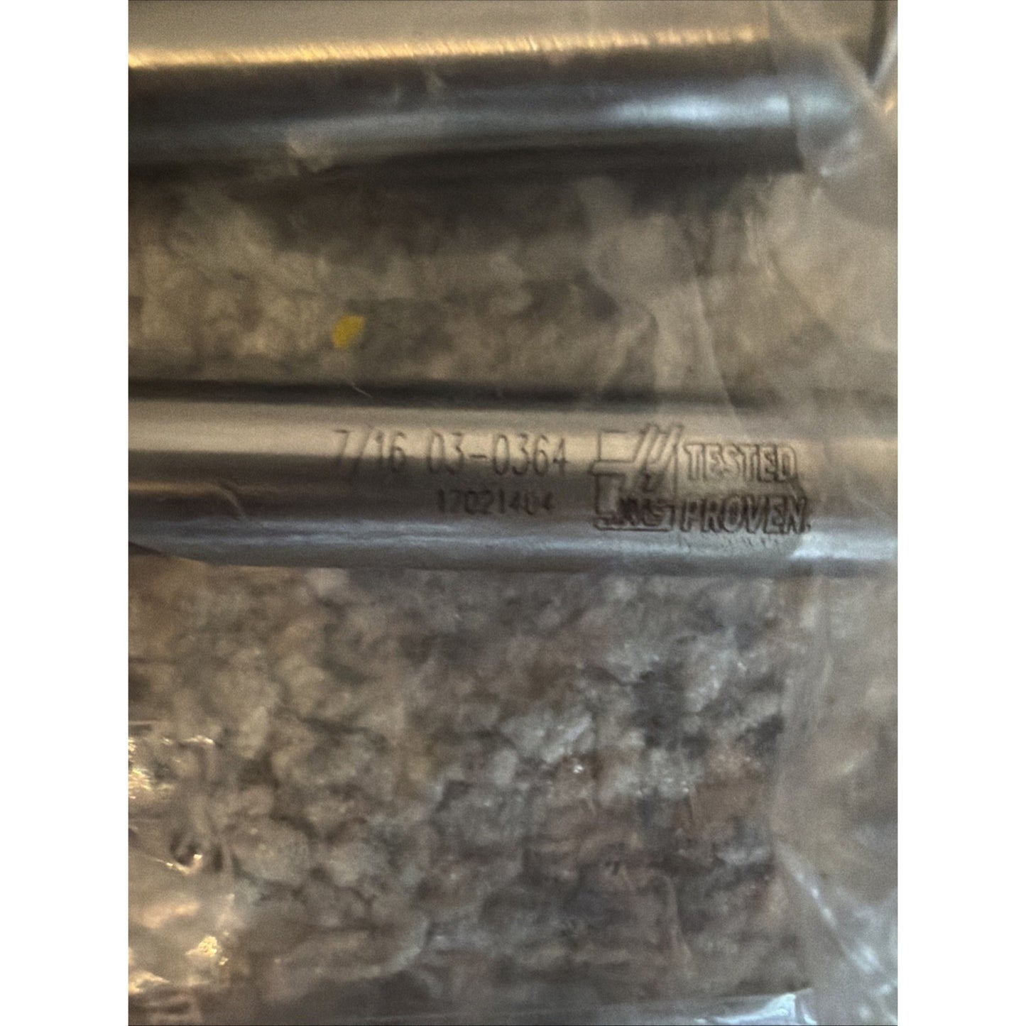 Close-up of a silver metal rod with engraved text including "TESTED PROVEN" and numerical codes, resting on a textured gray carpet inside clear plastic packaging.