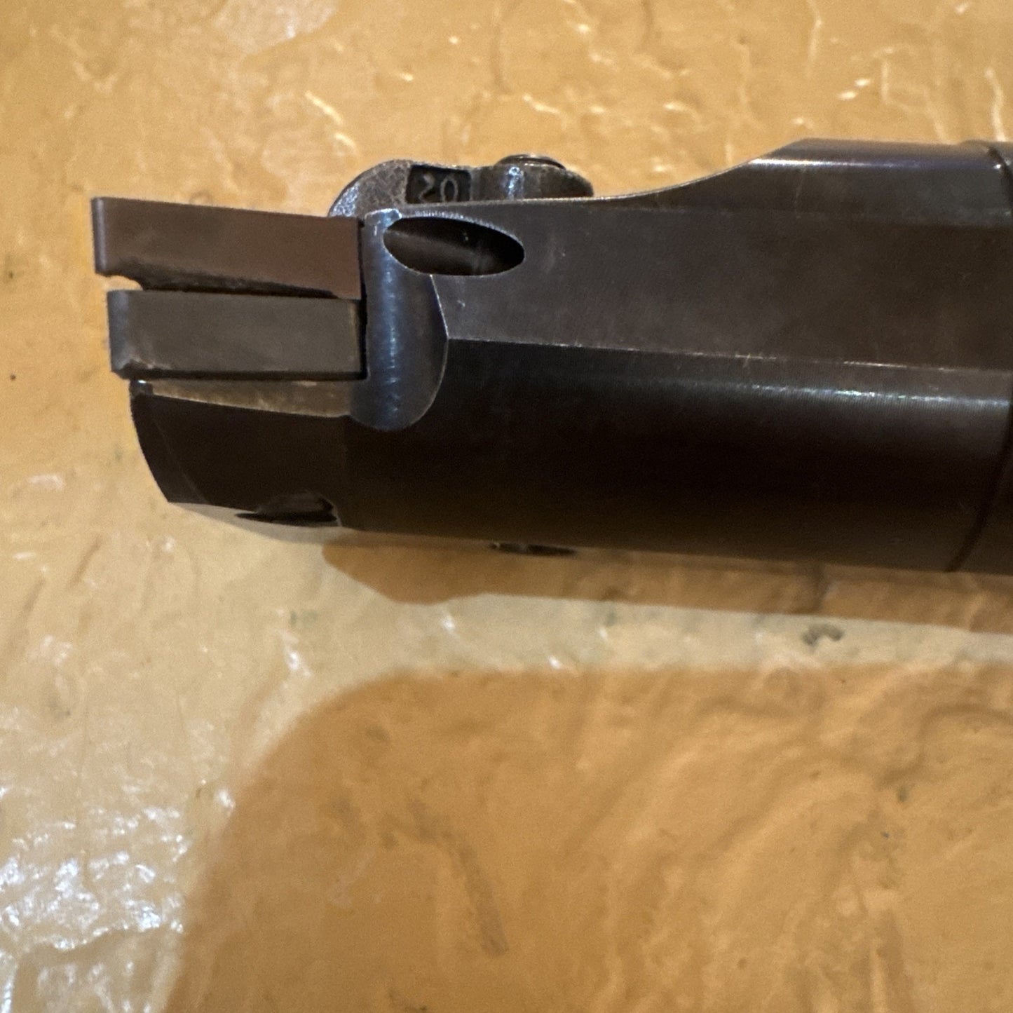 Close-up view of a dark metal cutting tool tip with two flat blades and a rounded notch near the base, resting on a textured beige surface.