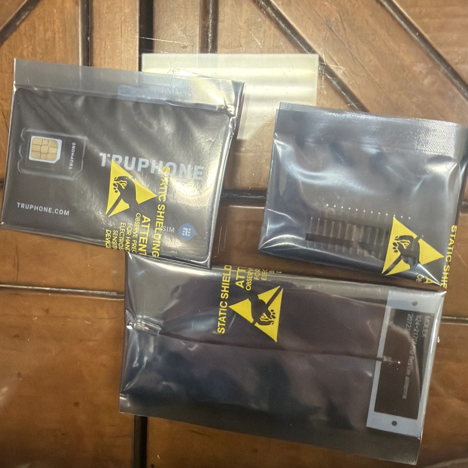 Three black antistatic static shielding bags containing electronic components, including a SIM card, a small circuit board, and a smartphone touchscreen digitizer, placed on a wooden surface.