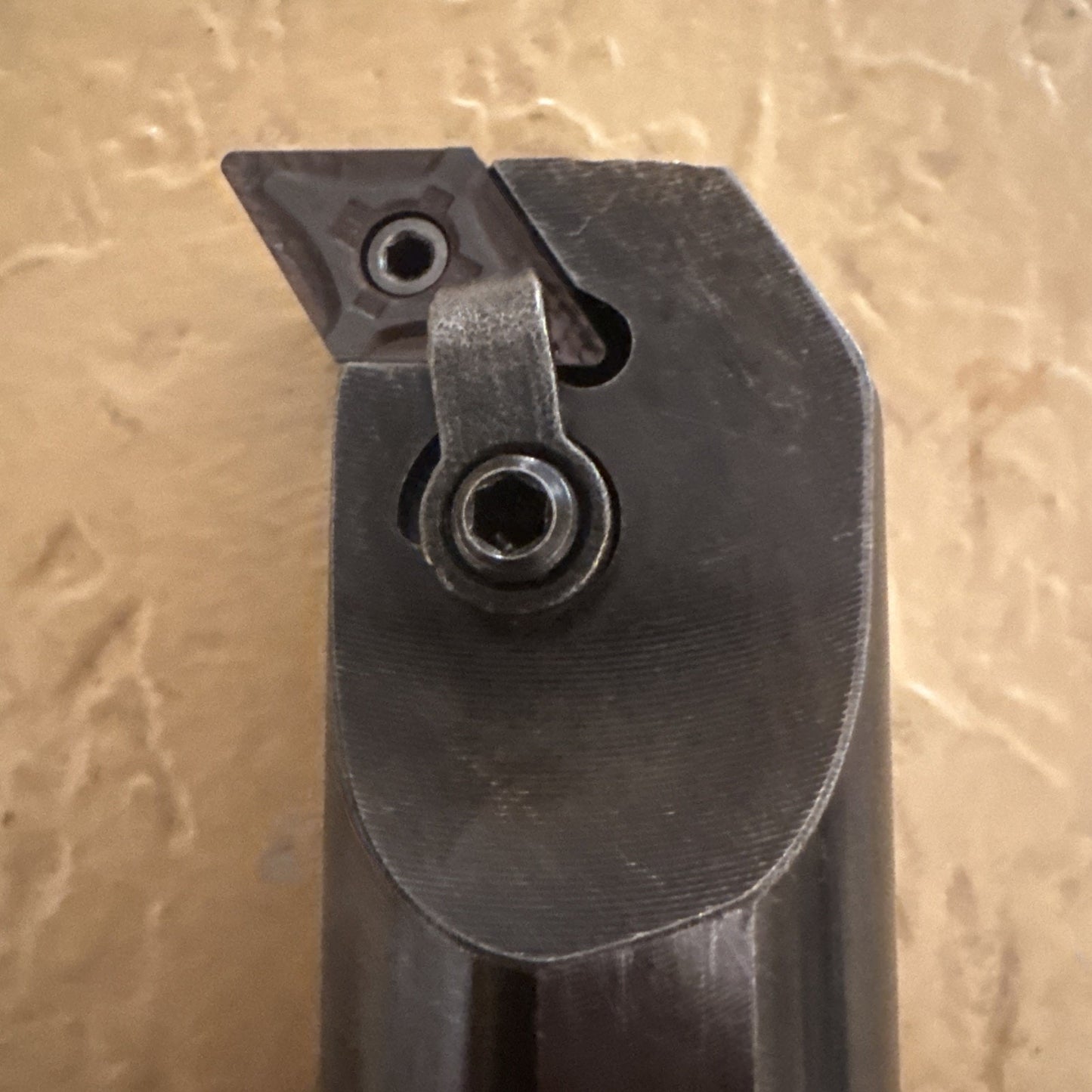 Close-up of a dark metal lathe cutting tool holder featuring a triangular carbide insert secured by a screw and clamp, set against a textured beige background.