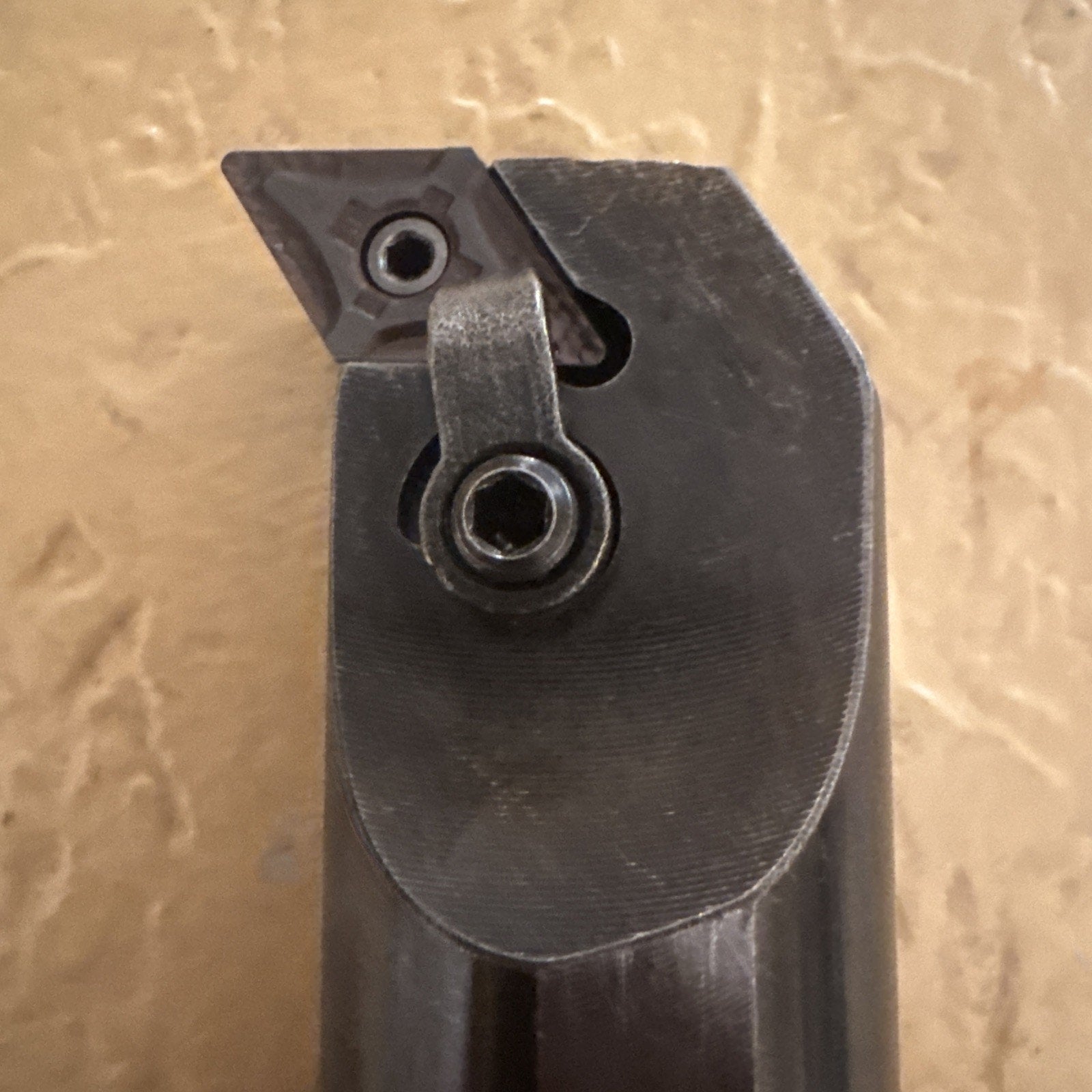 Close-up of a dark metal lathe cutting tool holder featuring a triangular carbide insert secured by a screw and clamp, set against a textured beige background.