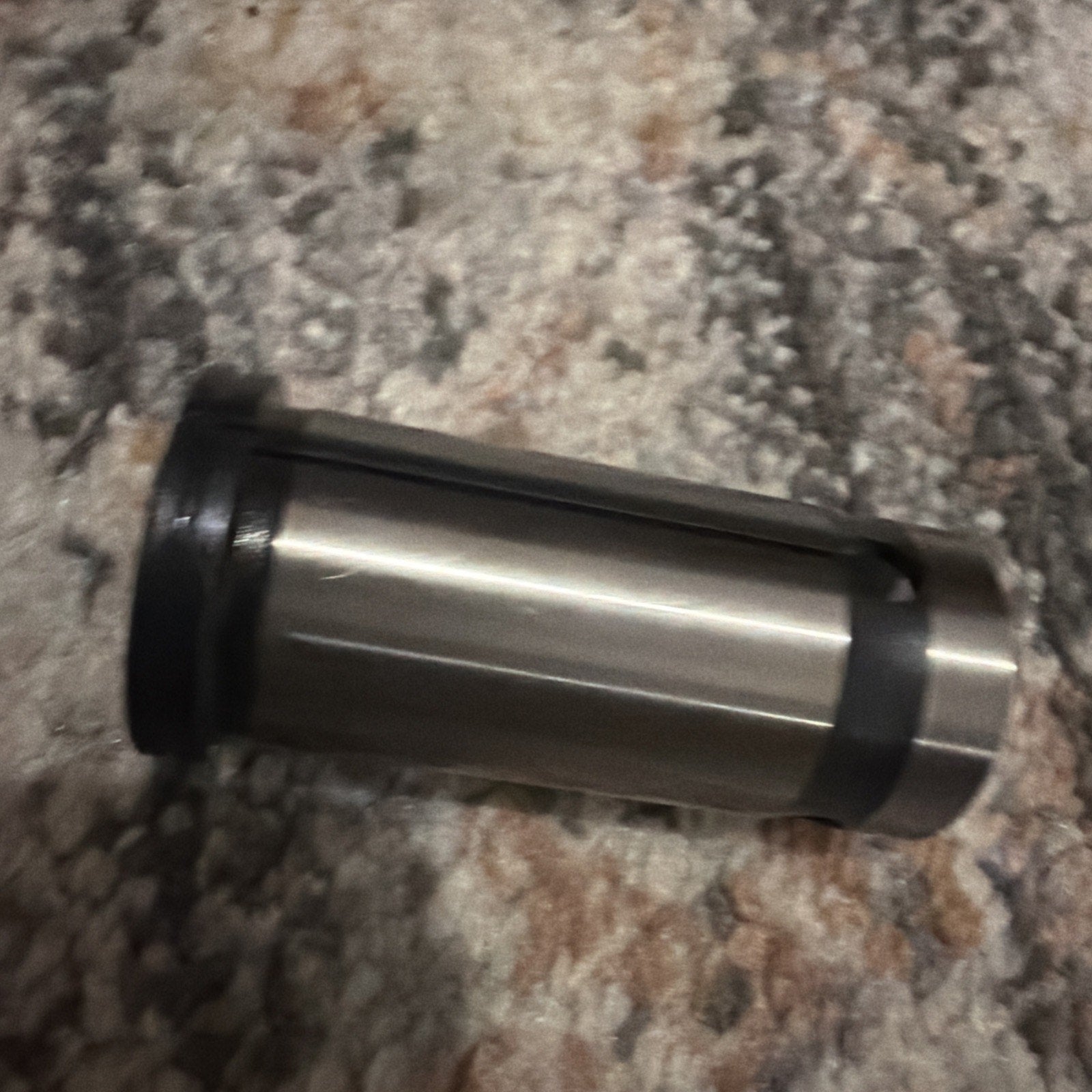 Metal cylindrical collet with three vertical slits and black rubber ends, lying horizontally on a multi-tone textured carpet surface.