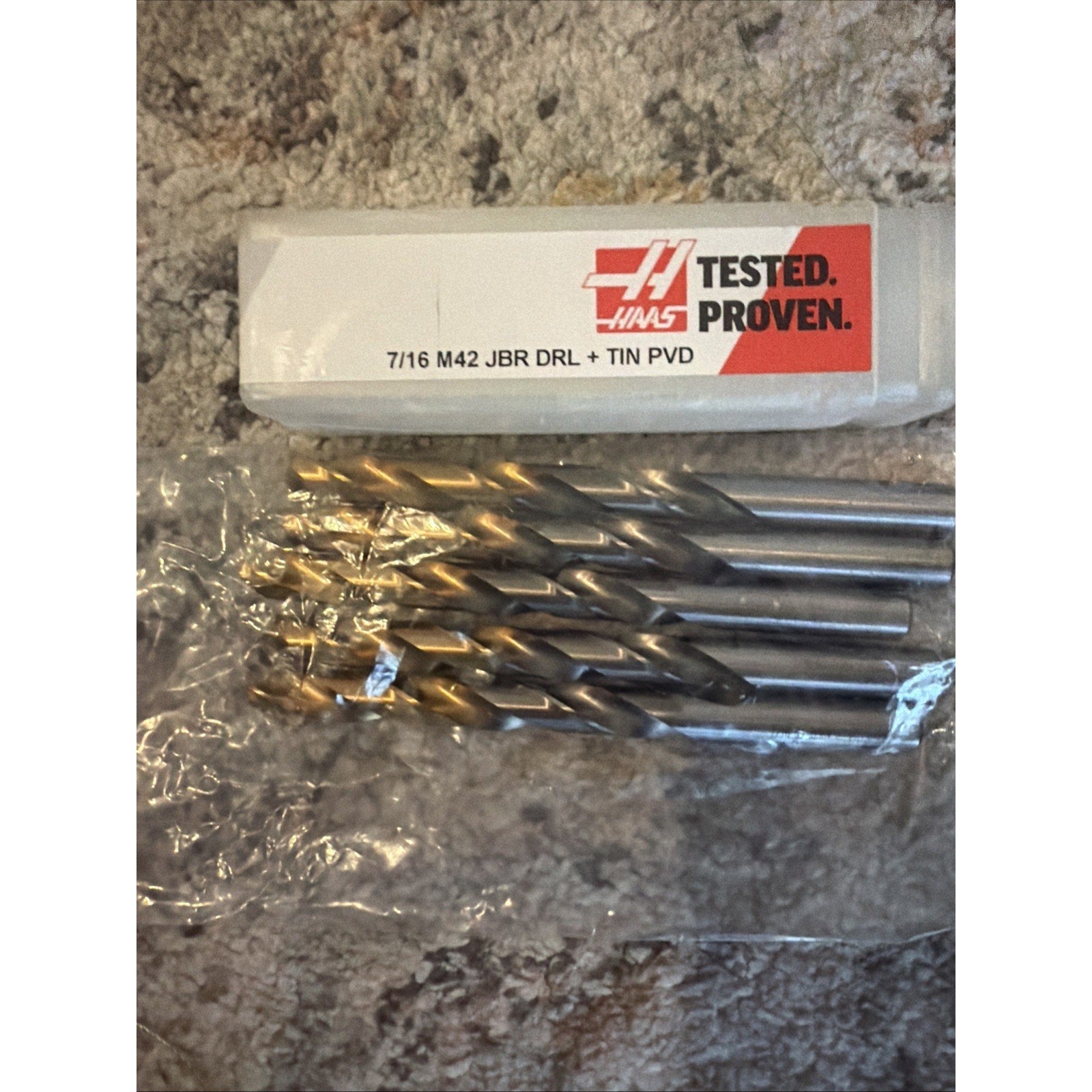 Set of five metal twist drill bits with titanium nitride coated tips partially wrapped in clear plastic, positioned beneath a white and red labeled plastic case on a textured gray surface.